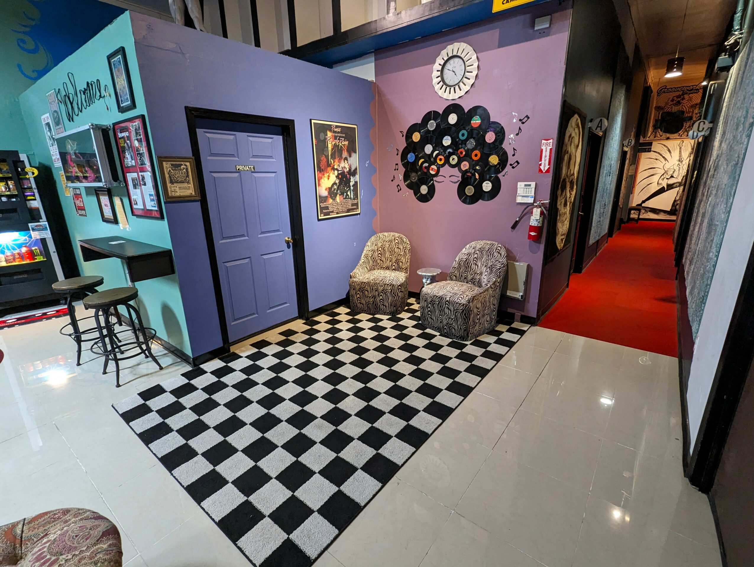 Corner with two chairs, checkered rug, purple and blue walls, clock, and wall art.