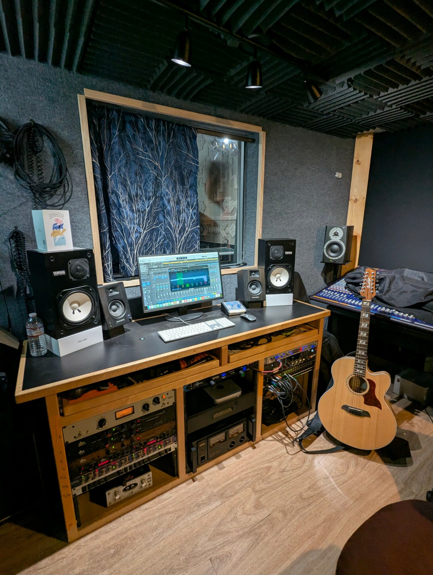 Music studio with mixing console, speakers, guitar, and soundproof window in a room with black ceiling and wooden floor.