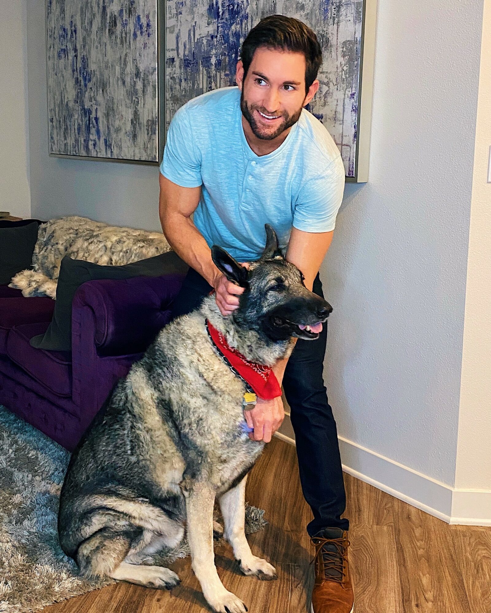 Man smiling with a large dog wearing a red bandana indoors, with artwork on the wall behind.