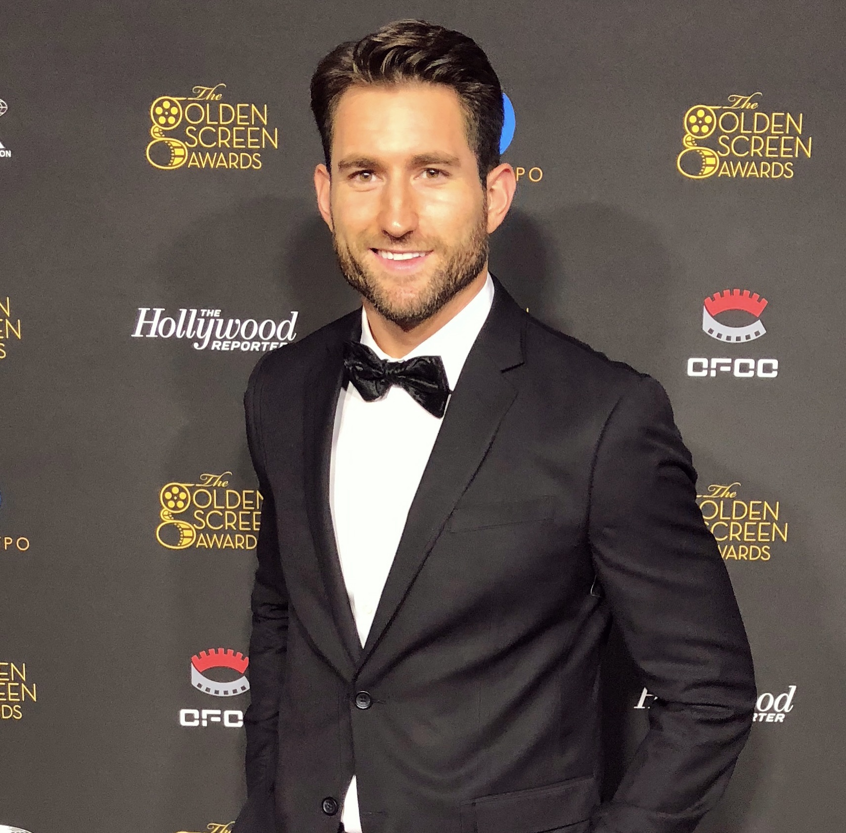 Man in tuxedo with bow tie smiling at event backdrop with logos.