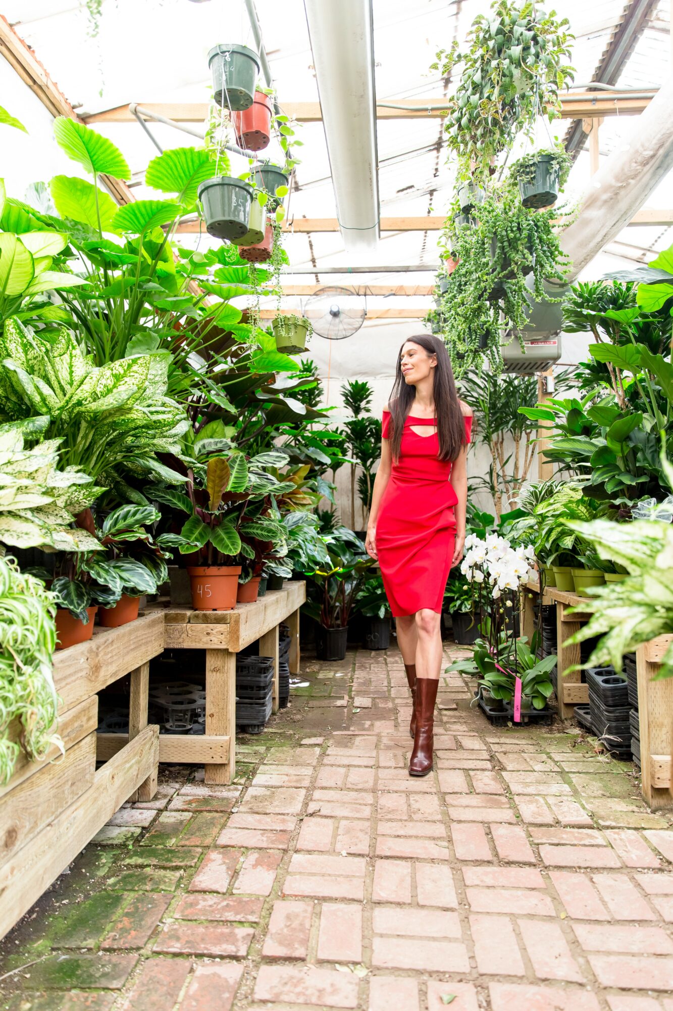 Woman in red dress walking through a greenhouse with lush green plants on both sides.