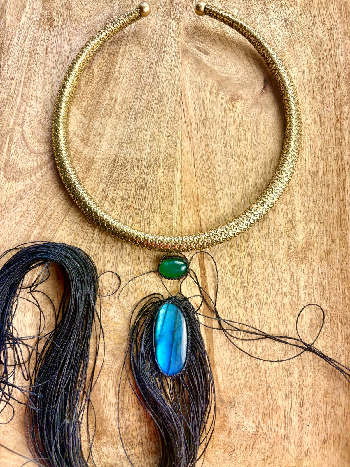 Gold-colored necklace with green and blue pendants on a wooden surface, with black thread and hair strands nearby.