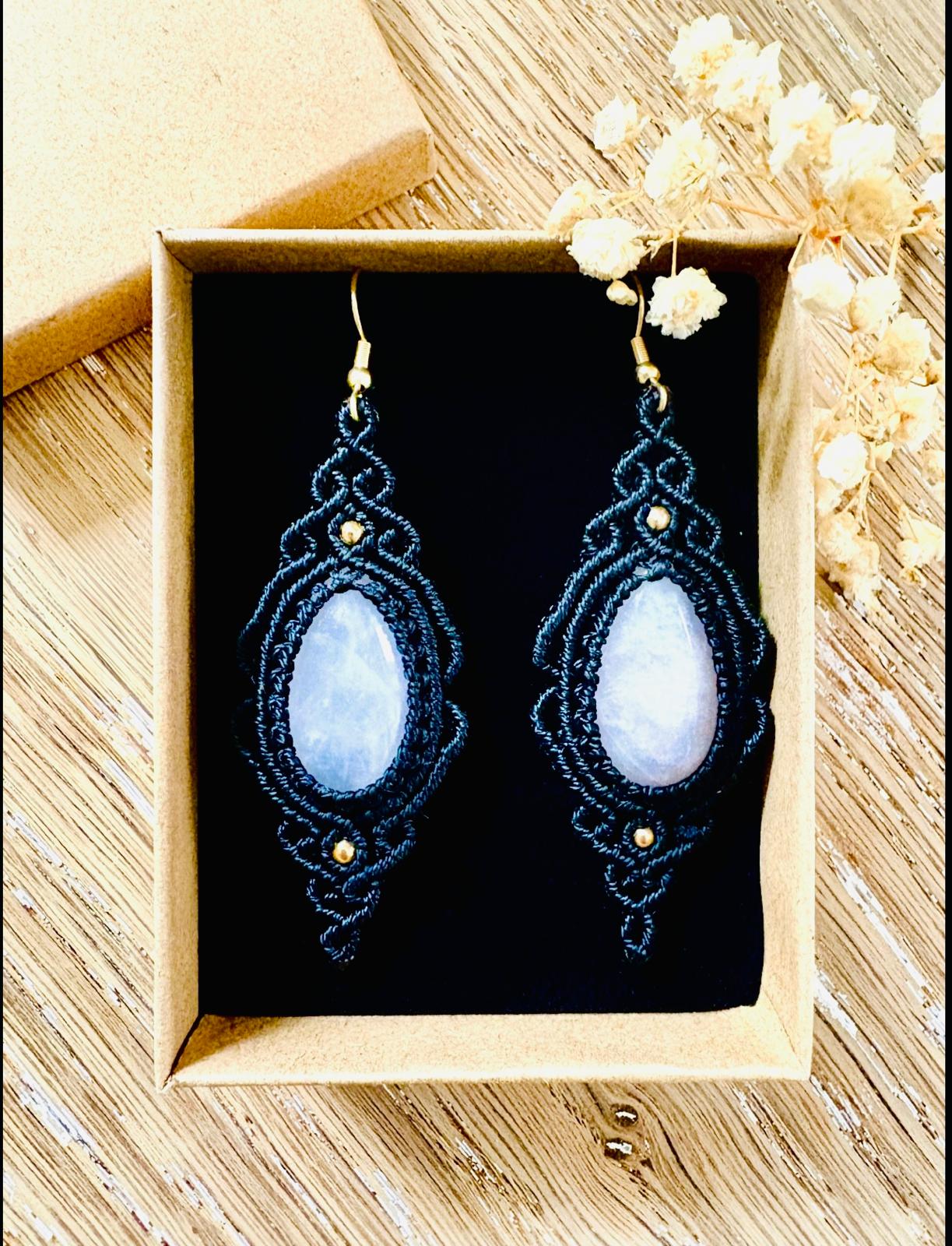 Pair of beaded earrings with oval stones in a box on a wooden surface, surrounded by dried flowers.