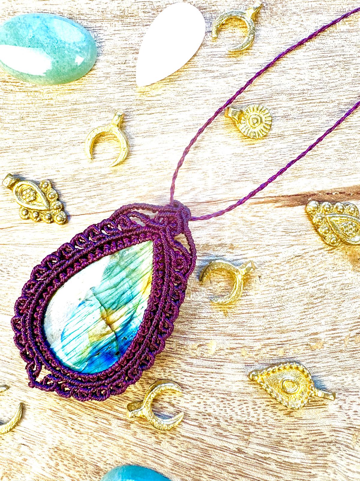 Purple macrame necklace with a large, colorful teardrop-shaped pendant, surrounded by small gold and shell charms on a wooden surface.