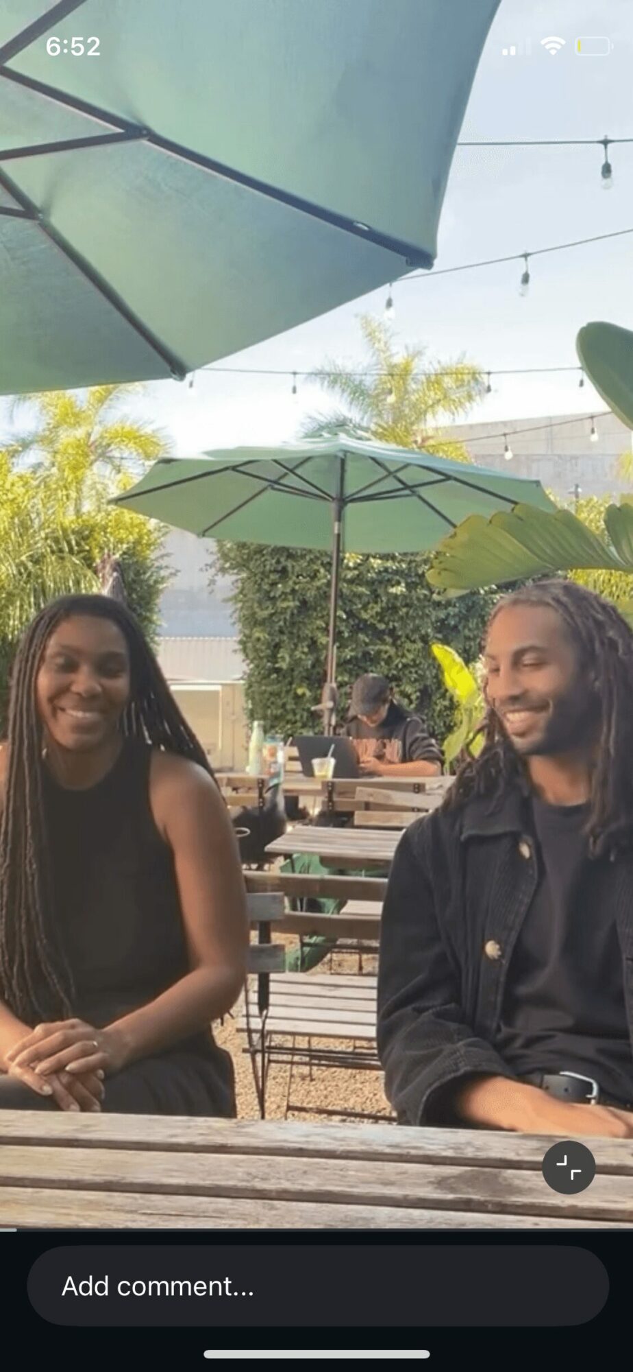 Two people sitting at a table outdoors under umbrellas, smiling, with trees and other tables in background.