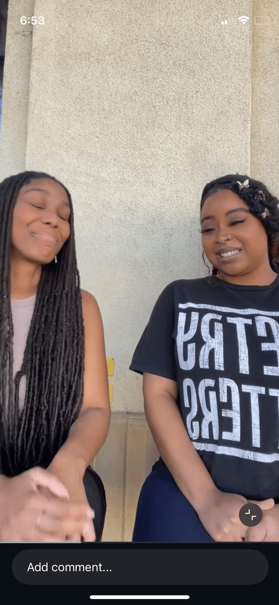 Two women sitting and smiling, one with long braids and the other with short curly hair, against a plain wall.