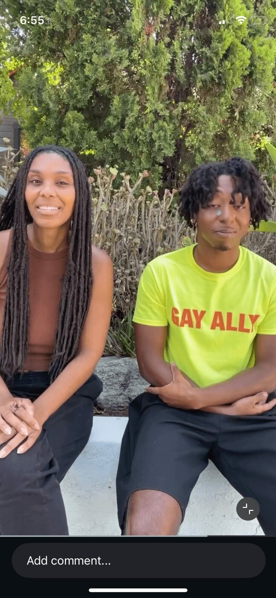 Two people sitting outdoors with trees in the background, smiling, one wearing a black top and the other a bright yellow shirt.