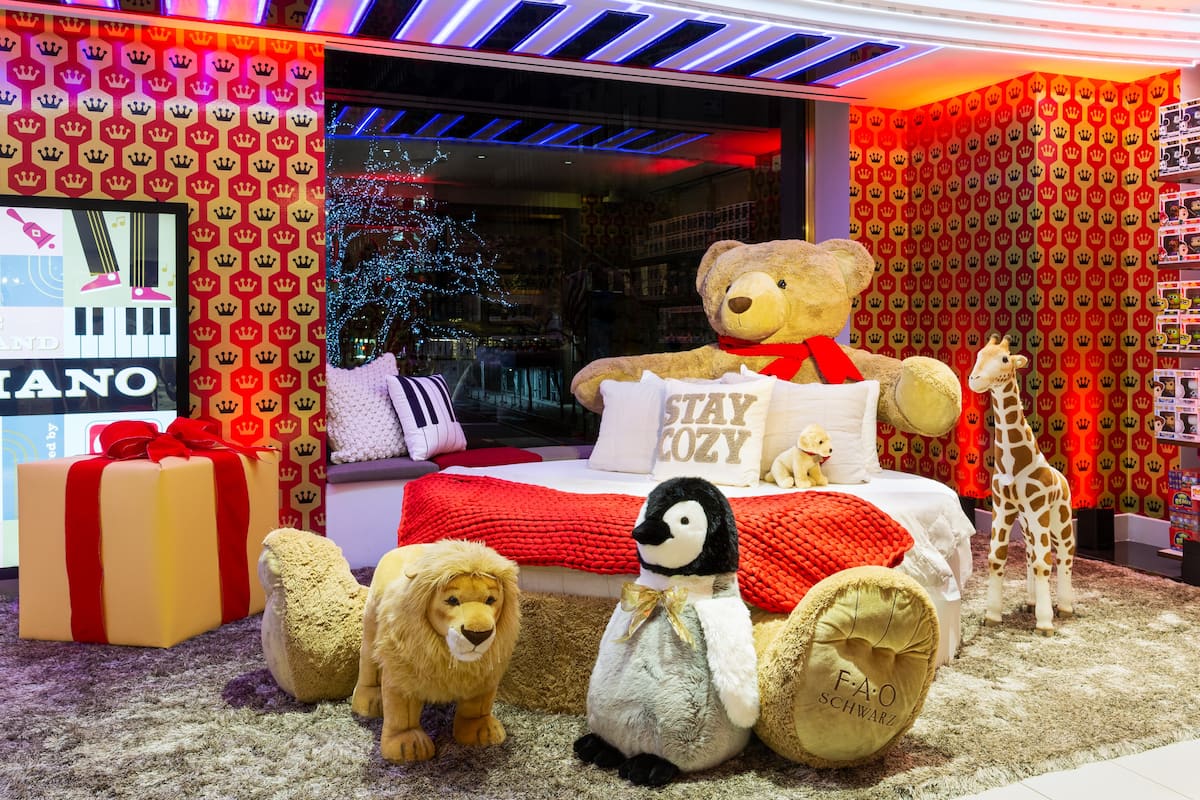 Room with teddy bears, a penguin plush, a giraffe, and gift boxes on a carpeted floor, decorated with colorful wallpaper and lighting.