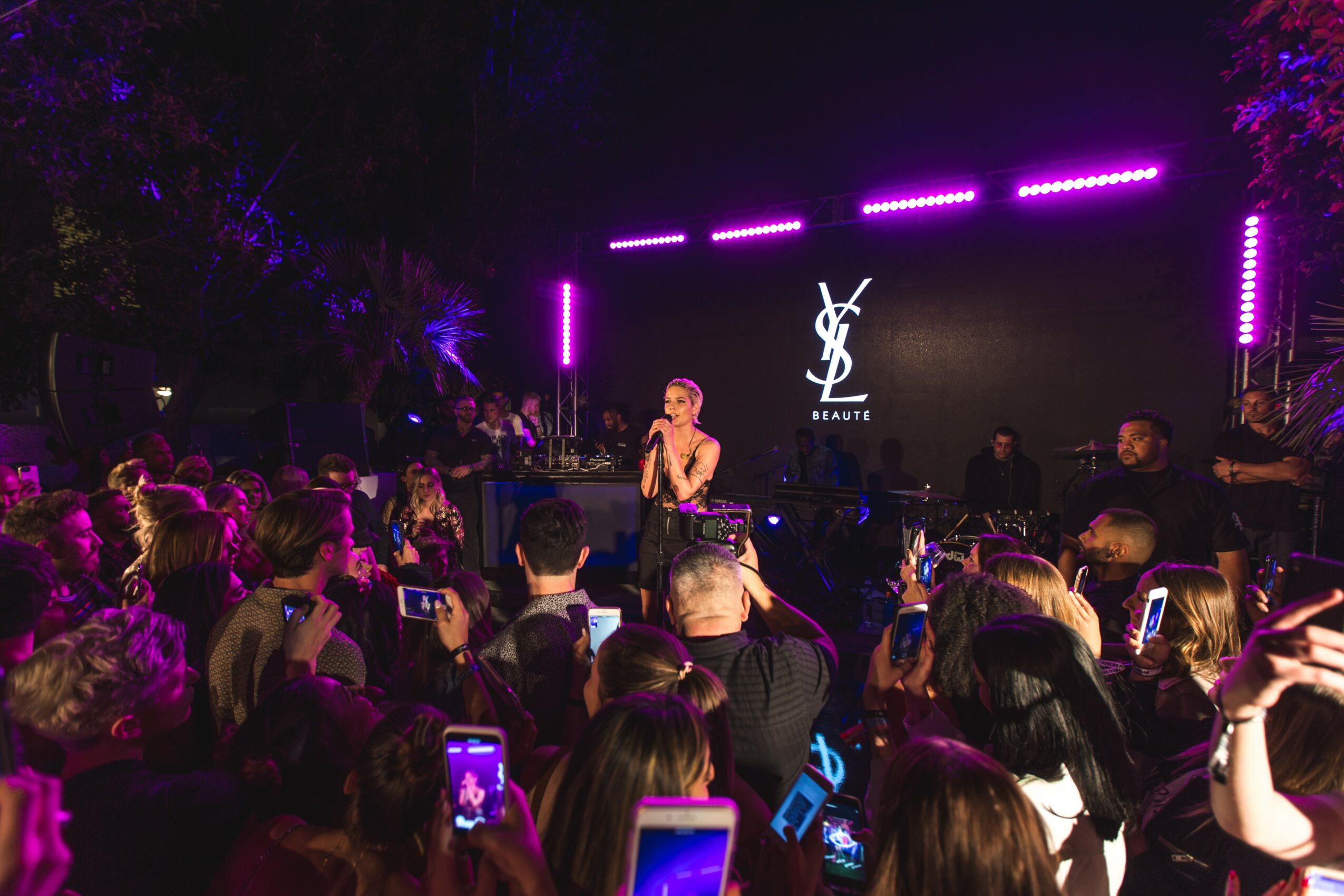 Performer on stage with YSL logo, audience watching at night with purple lighting, some taking photos.