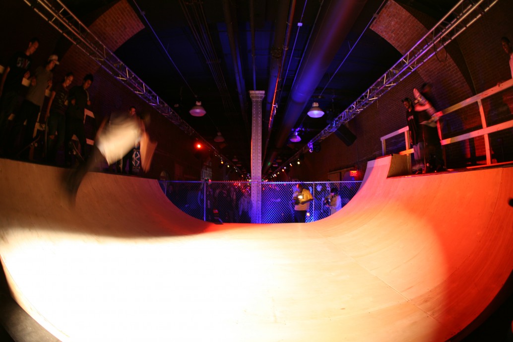 Indoor skateboarding ramp with people watching, illuminated by colorful lights, and a central pole in the middle.