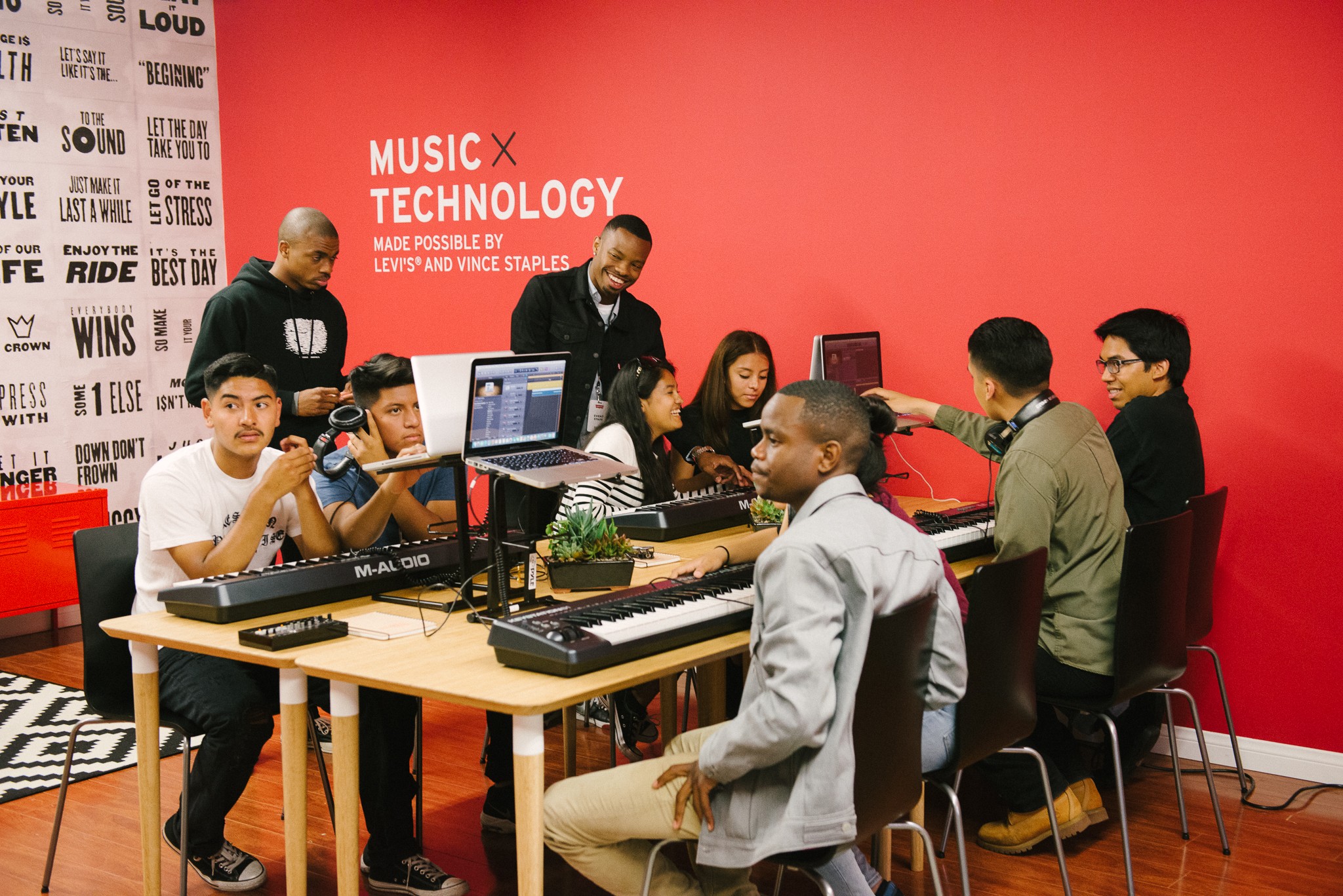 Group of people in a music technology workshop with computers and equipment on a red wall background.