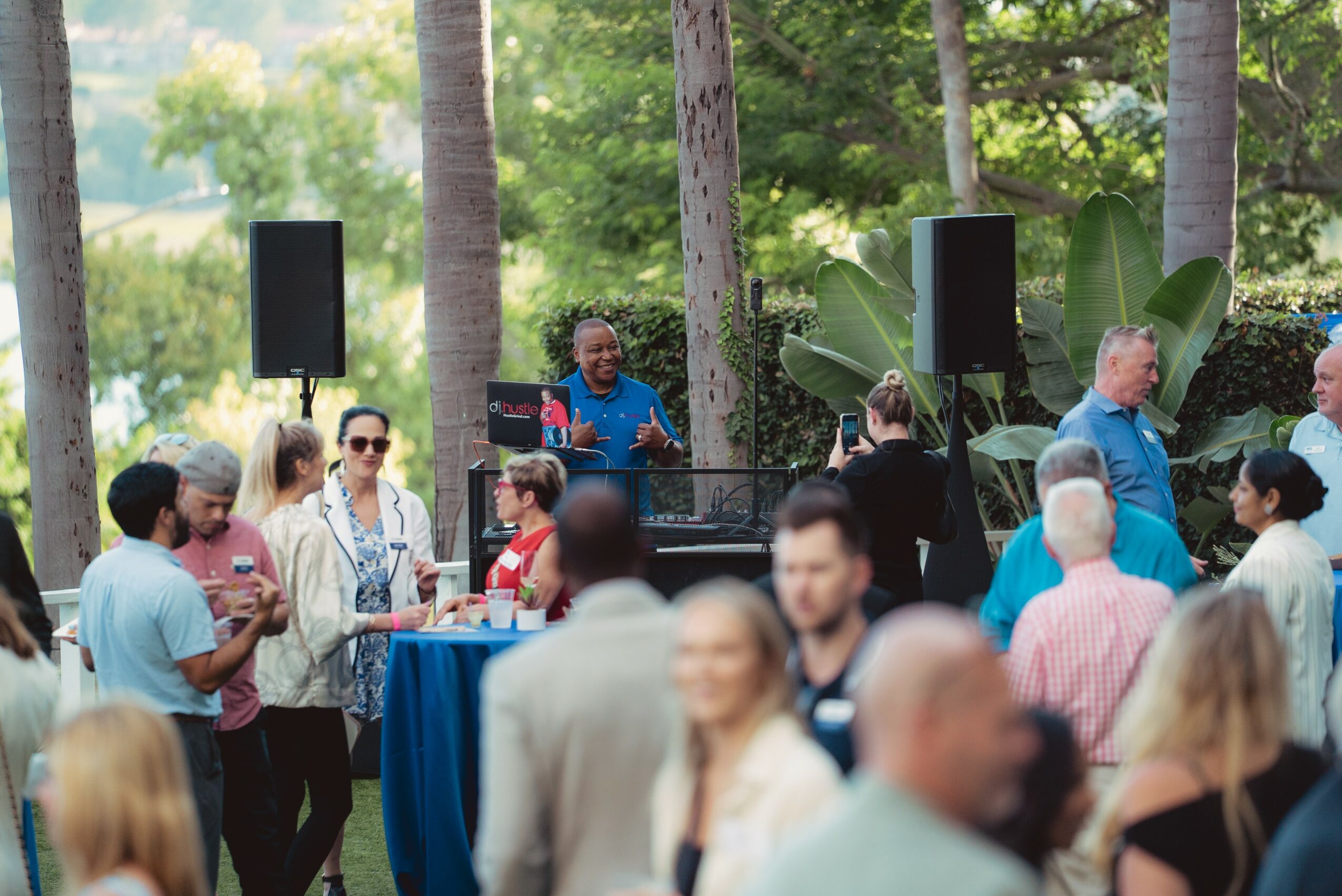 People gathered outdoors around a DJ with speakers, trees and greenery in the background, some holding drinks and talking.