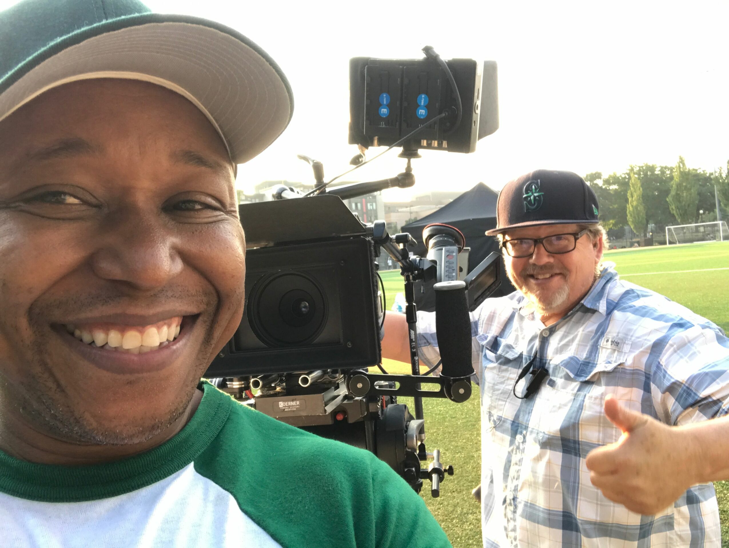 Two men outdoors near camera equipment, smiling, one giving a thumbs-up, field and trees in background.