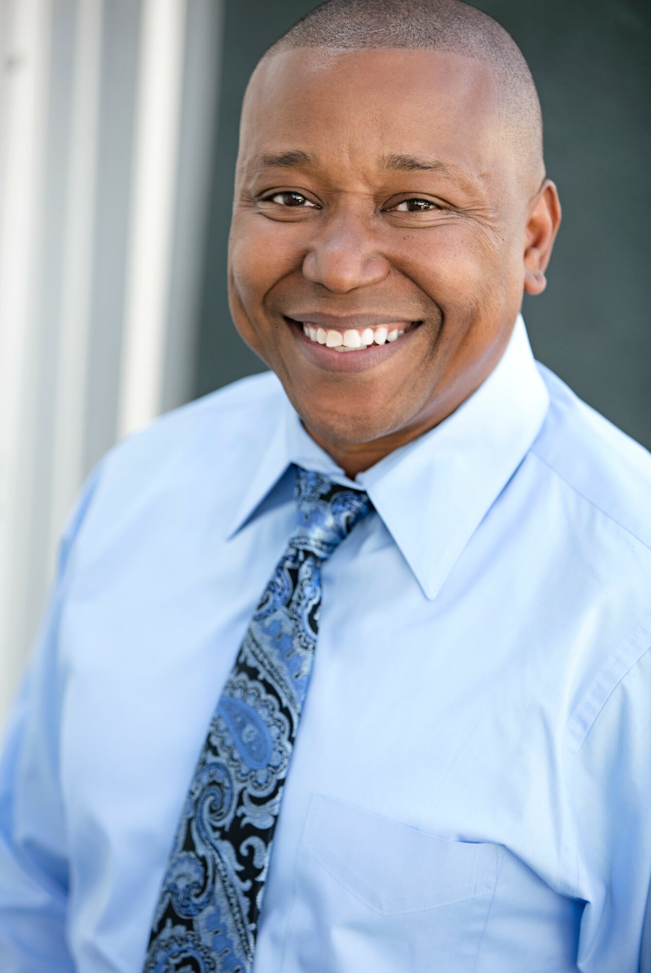 Smiling man in light blue shirt and patterned tie outdoors, with a building in the background.