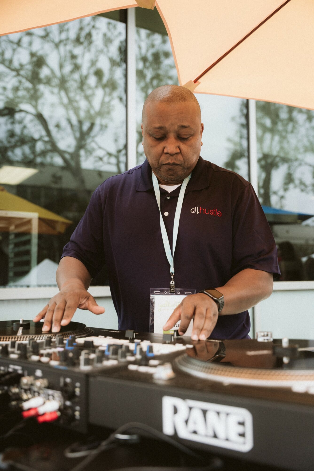 Person DJing outdoors under a large umbrella, with trees and buildings in the background.