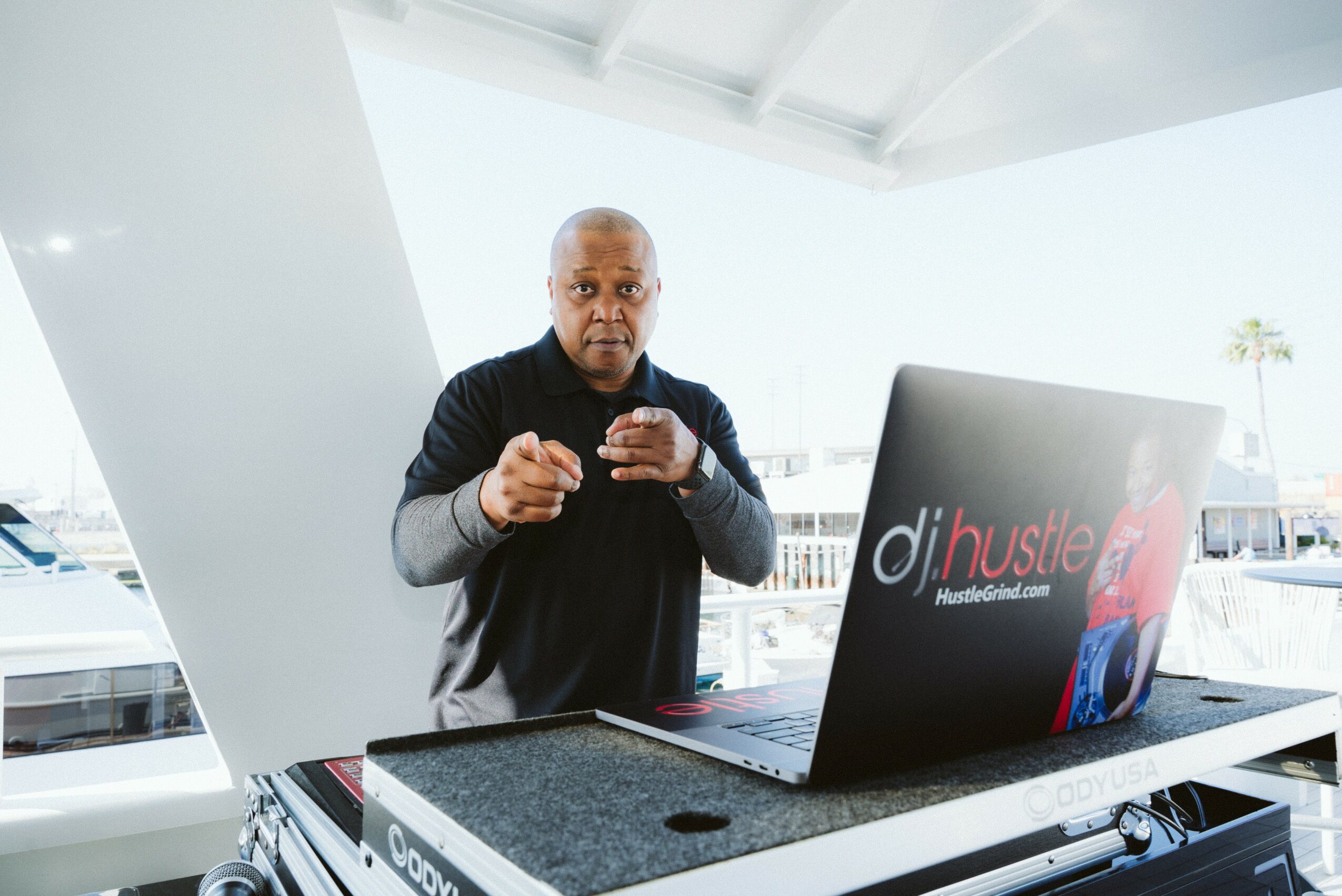 Man standing behind a table with a laptop outdoors, pointing at the camera, with a marina and boats in the background.