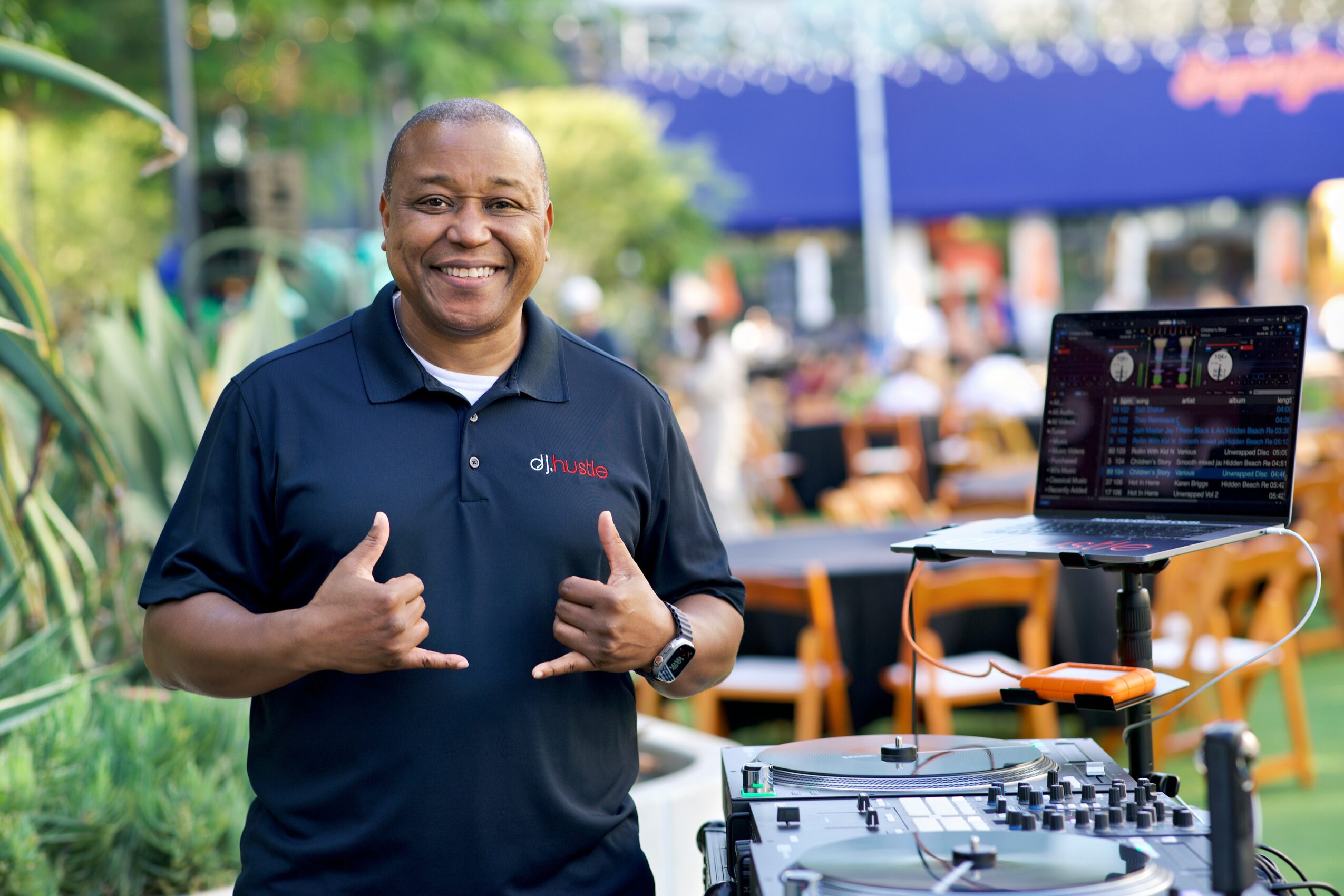 Smiling man in navy polo shirt standing outdoors with DJ equipment and a laptop nearby.