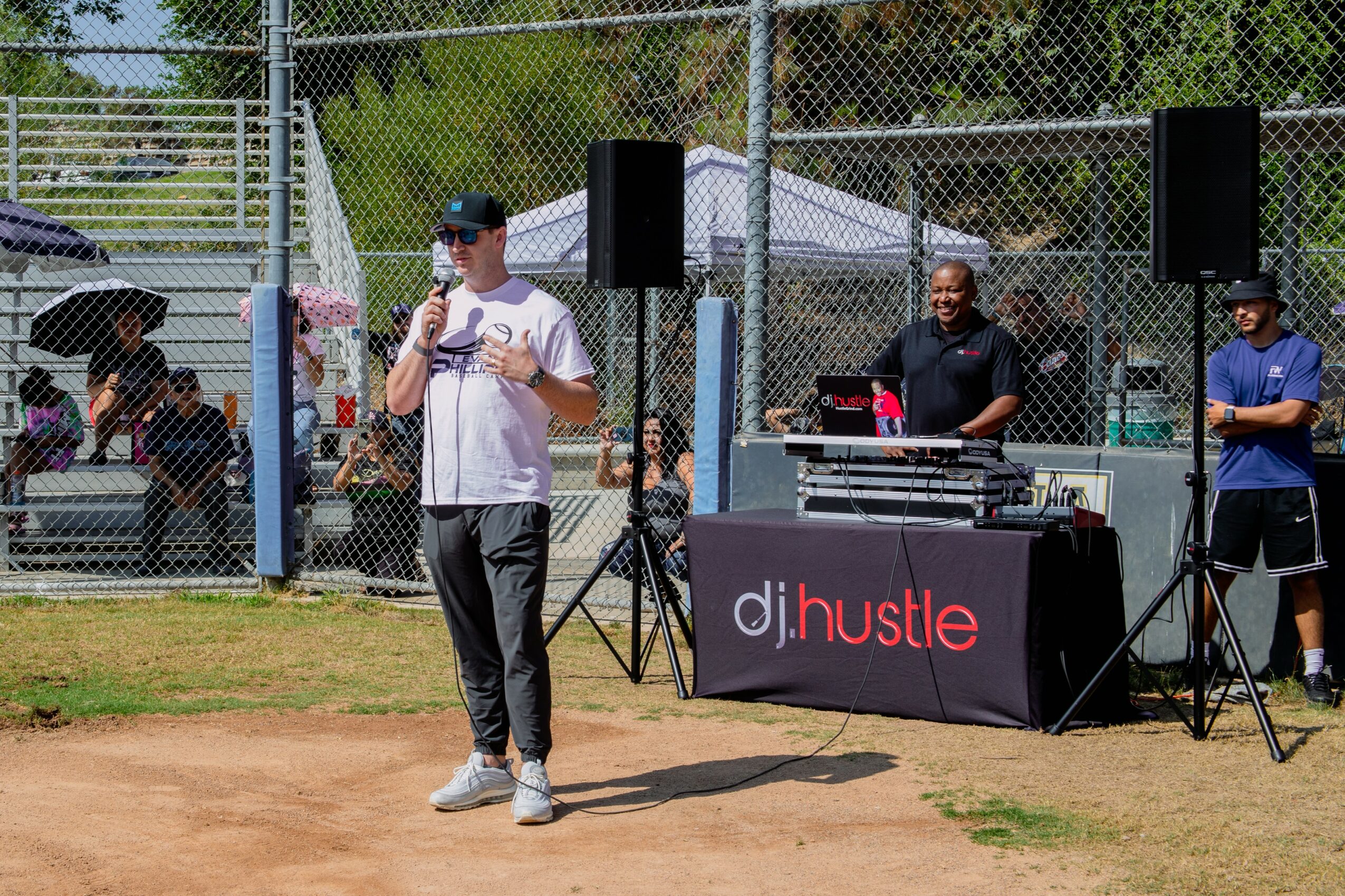 Person speaking into microphone at outdoor event with DJ setup and two people behind table, fenced sports field in background.