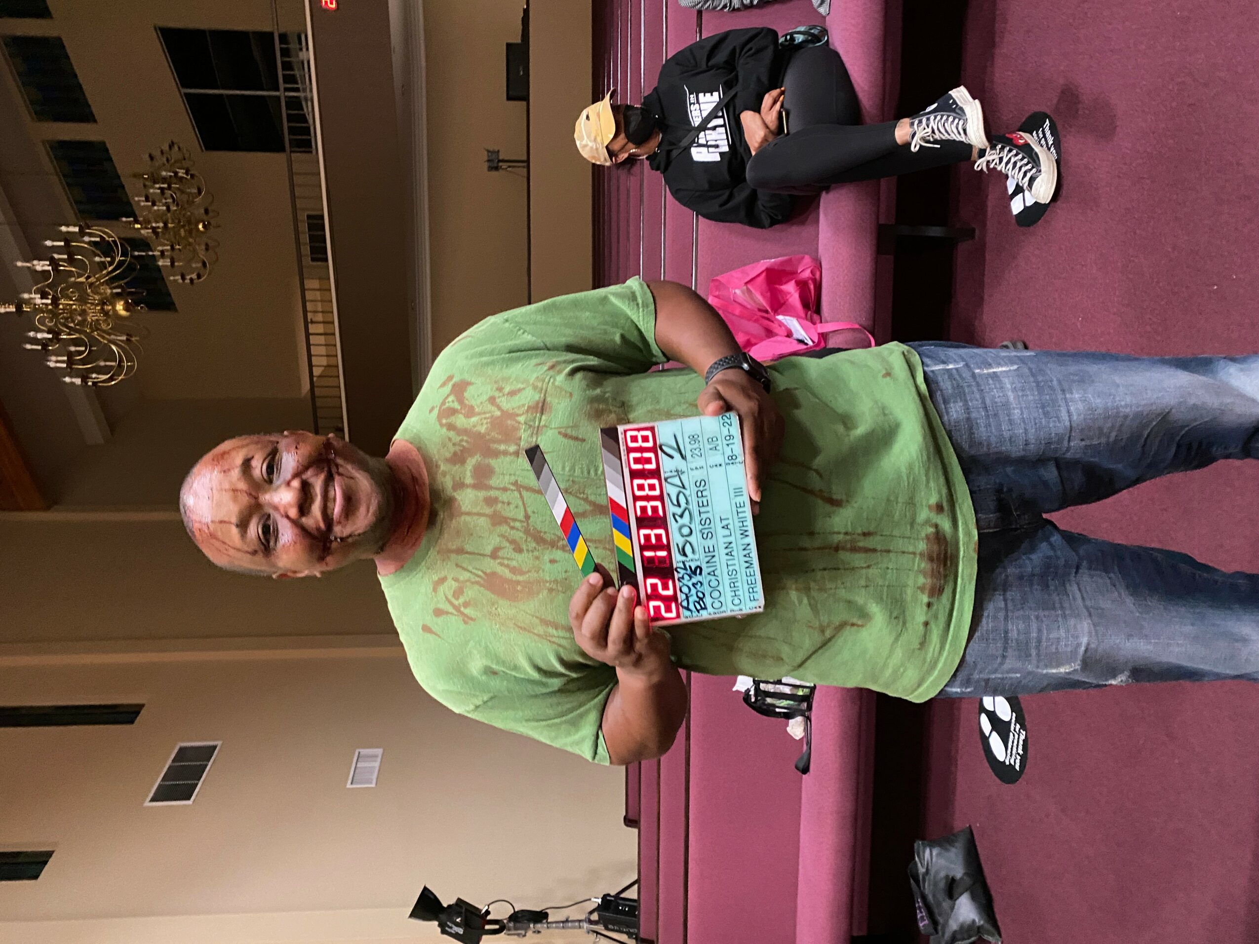 Man in green shirt holding a clapperboard, standing in a room with pink carpet and people in the background.