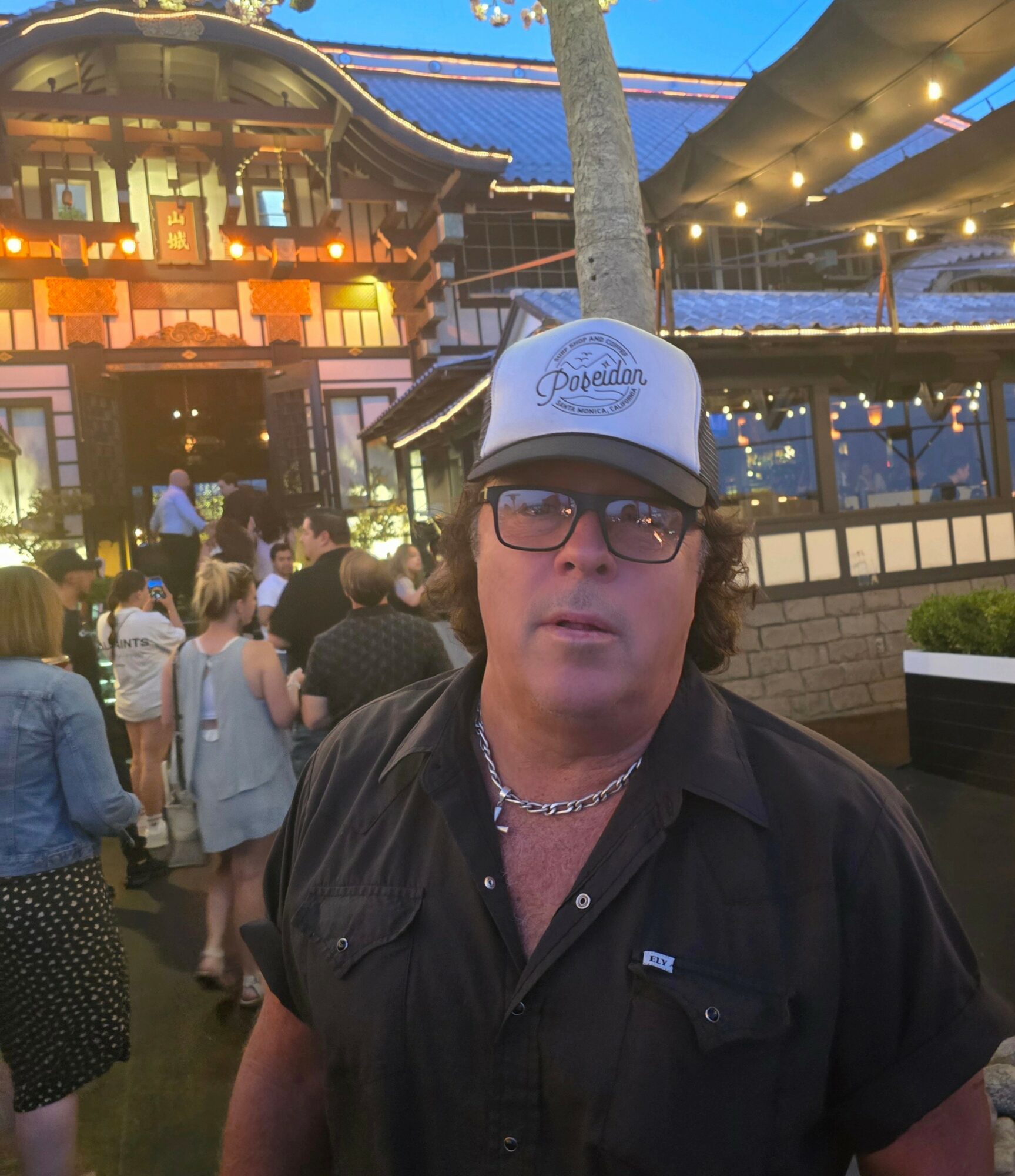Man wearing glasses and a trucker hat with a crowd and building in the background.
