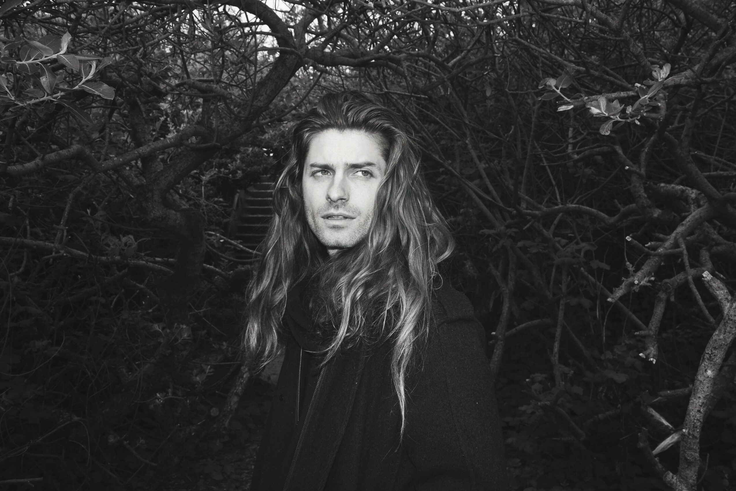 Woman with long hair standing outdoors among dark, twisted tree branches.