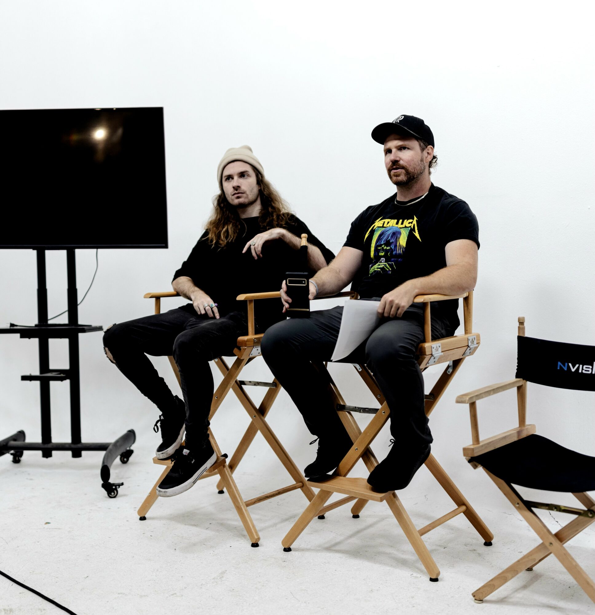 Two men sit on director's chairs in a white room, one with a beard and cap, the other with long hair and beanie.