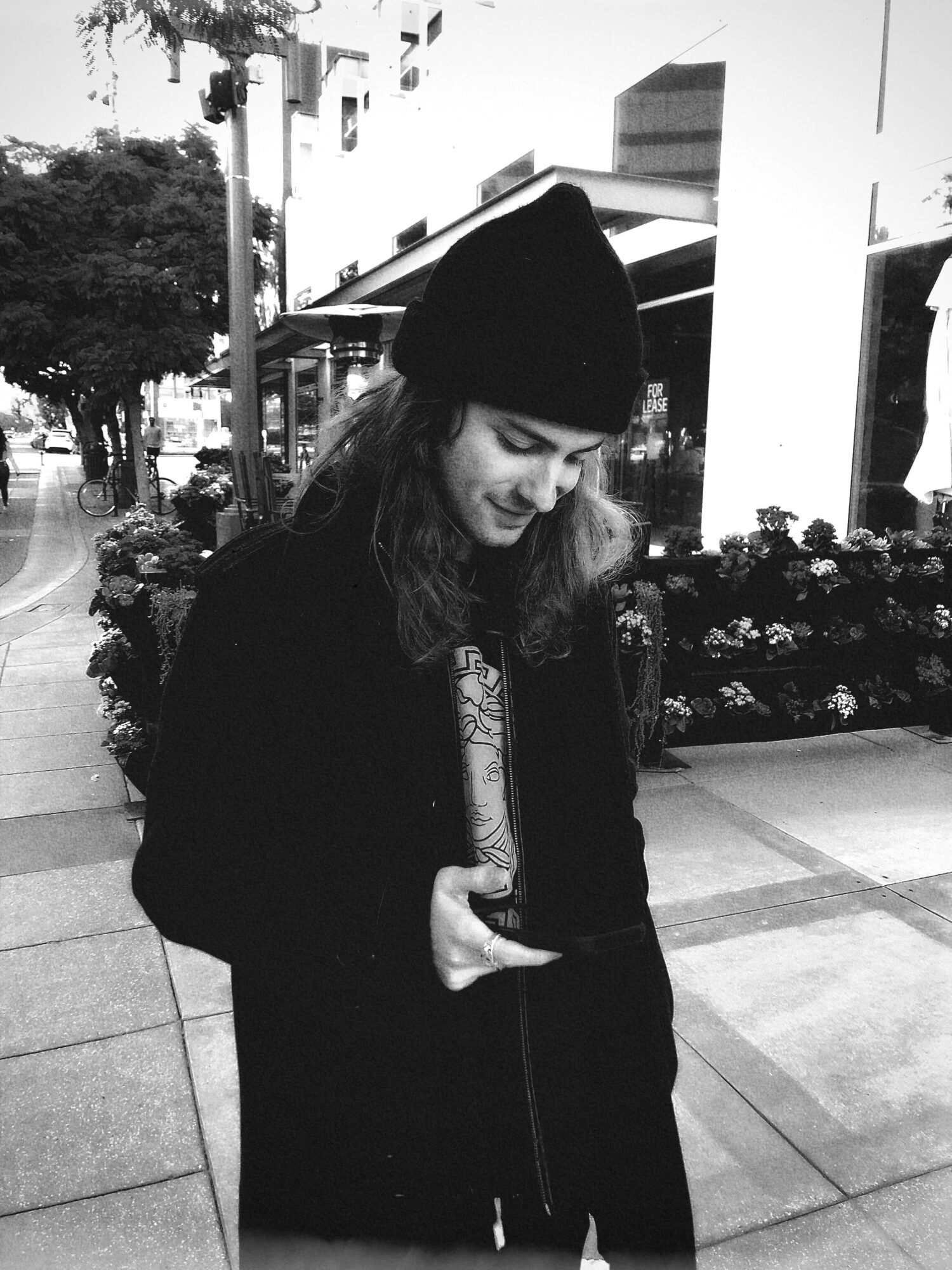 Young woman in a beanie looking at her phone outdoors, with buildings and trees in the background.