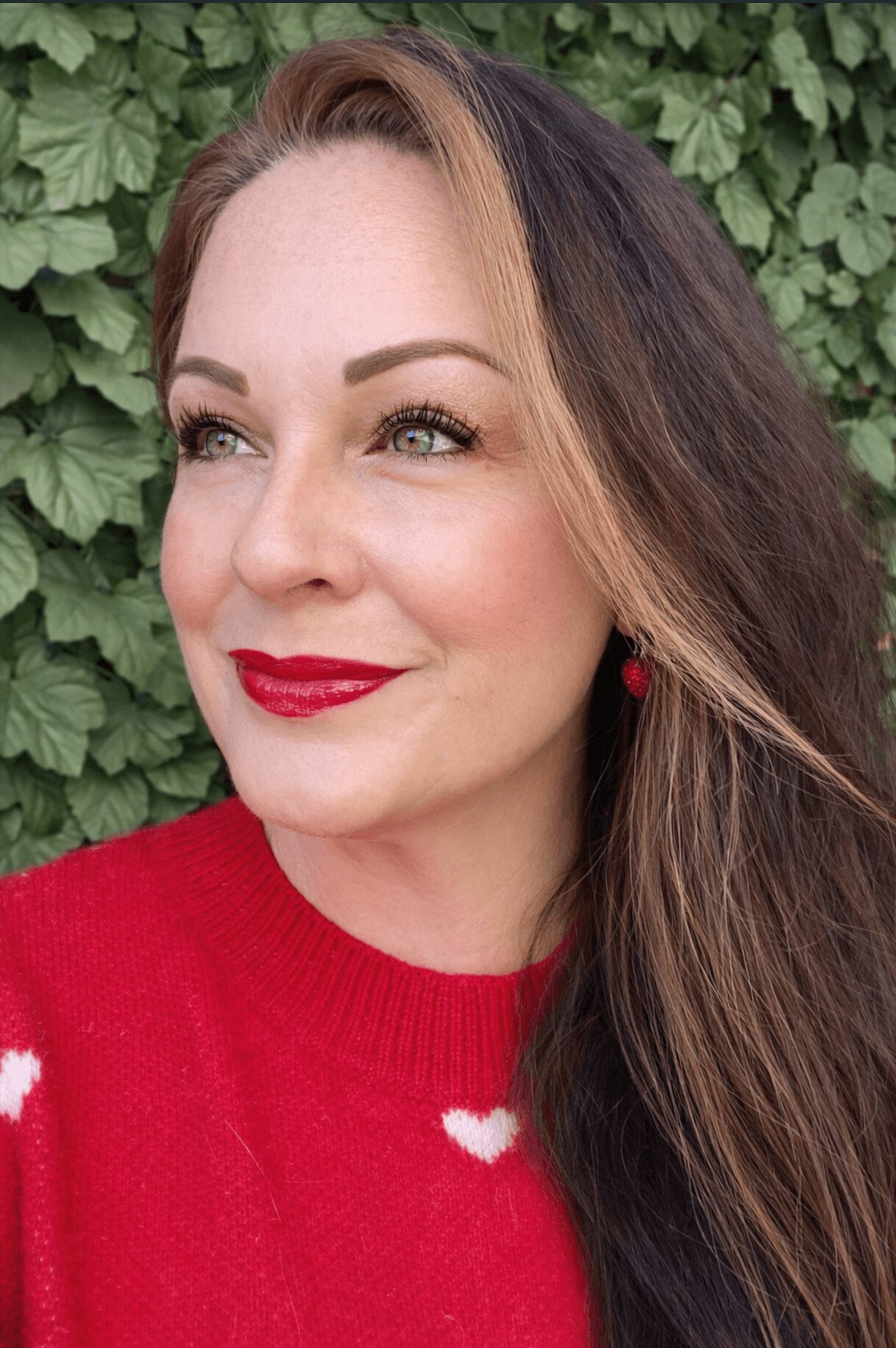 Woman with long brown hair, red lipstick, wearing a red sweater with white heart details, standing outdoors with green foliage background.