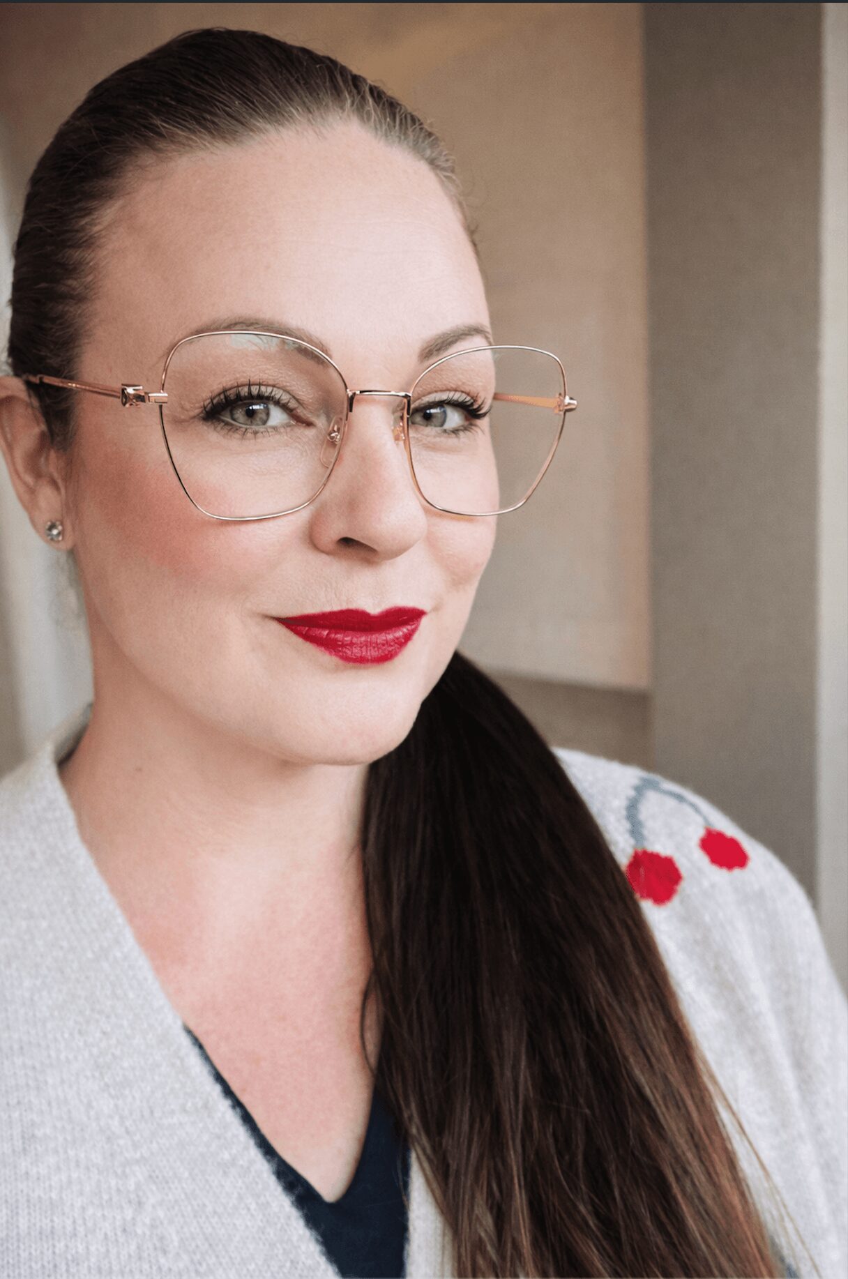 Woman with glasses, red lipstick, dark hair, wearing a light sweater with red heart patterns.