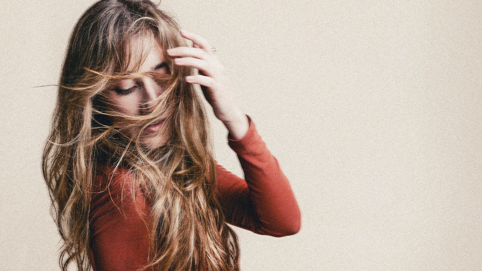 Woman with long hair touching her forehead, wearing a red long-sleeve top, against a plain background.