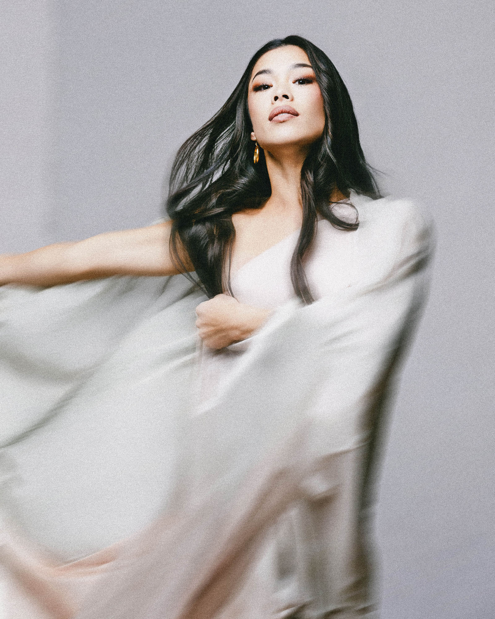 Woman with long dark hair wearing a flowing white garment, posing against a plain gray background.