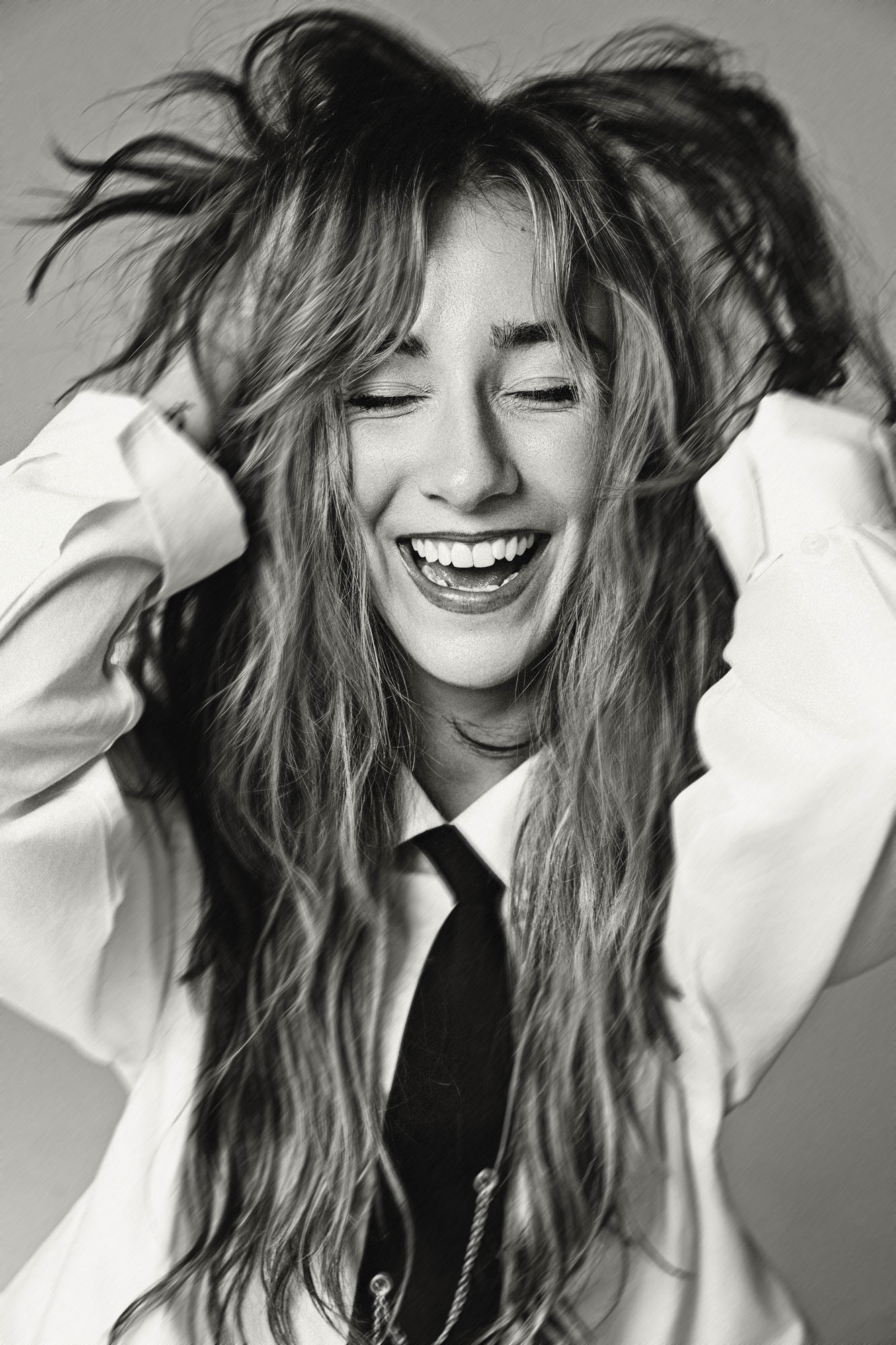 Woman with long, messy hair smiling and holding her head with both hands, wearing a white shirt and tie.