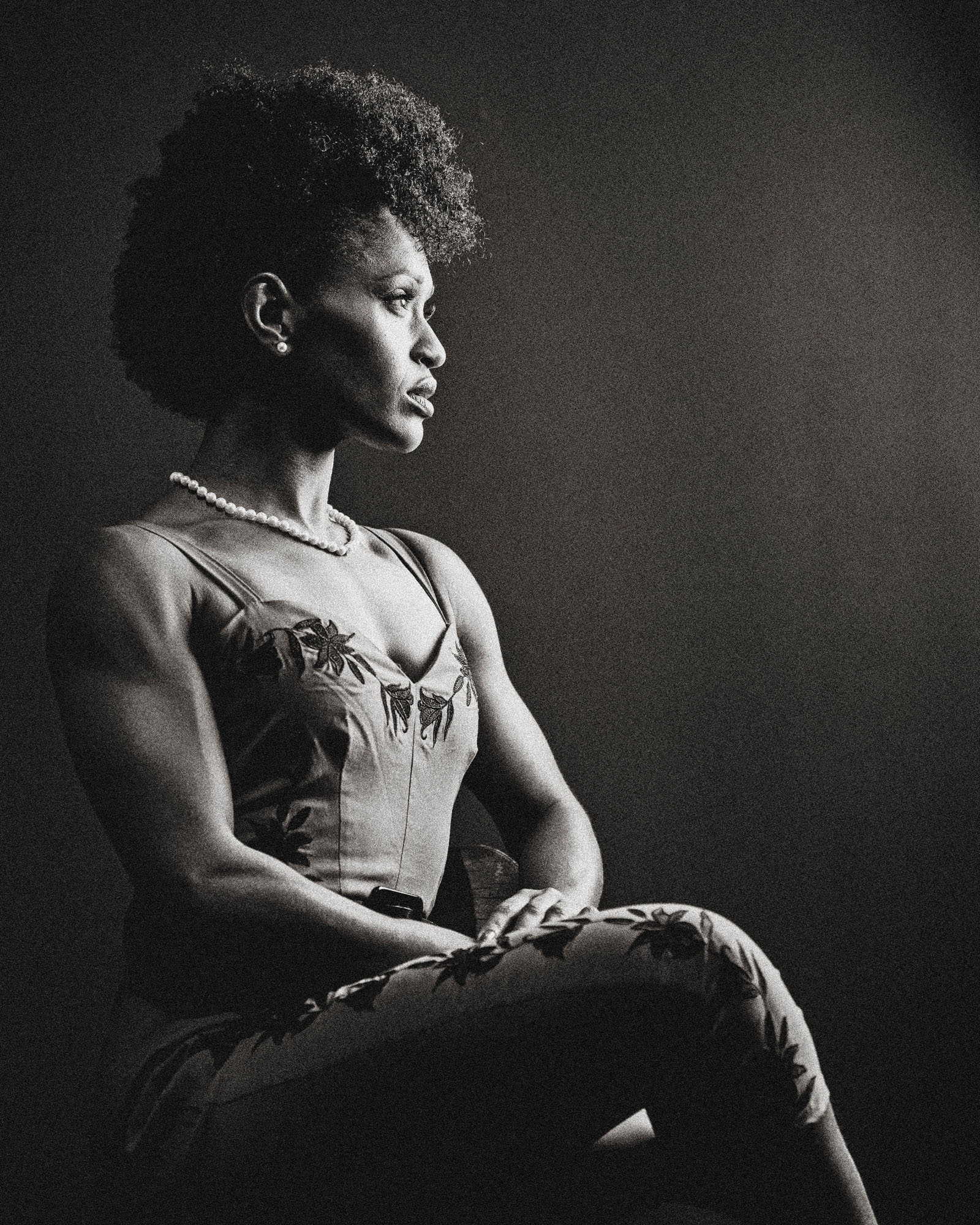 Side profile of a woman with natural hair, sitting with crossed arms, wearing a sleeveless top and pants.