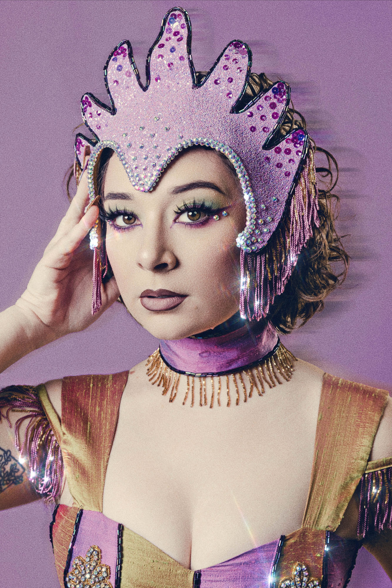 Person wearing a glittery crown with purple and pink accents, colorful costume, and makeup, against a purple background.