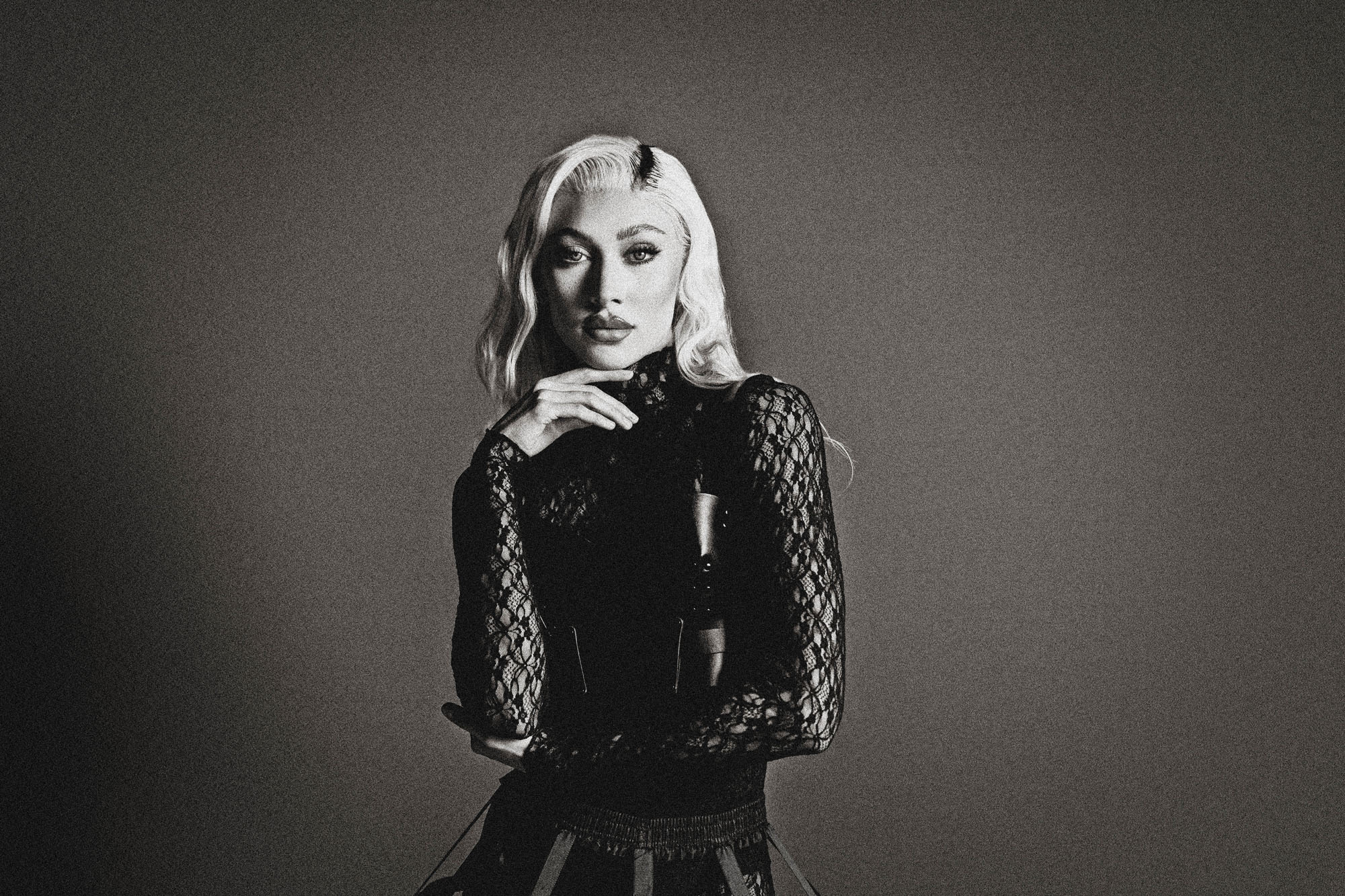 Woman with wavy hair wearing a lace long-sleeve top, posing with hand near chin against plain background.