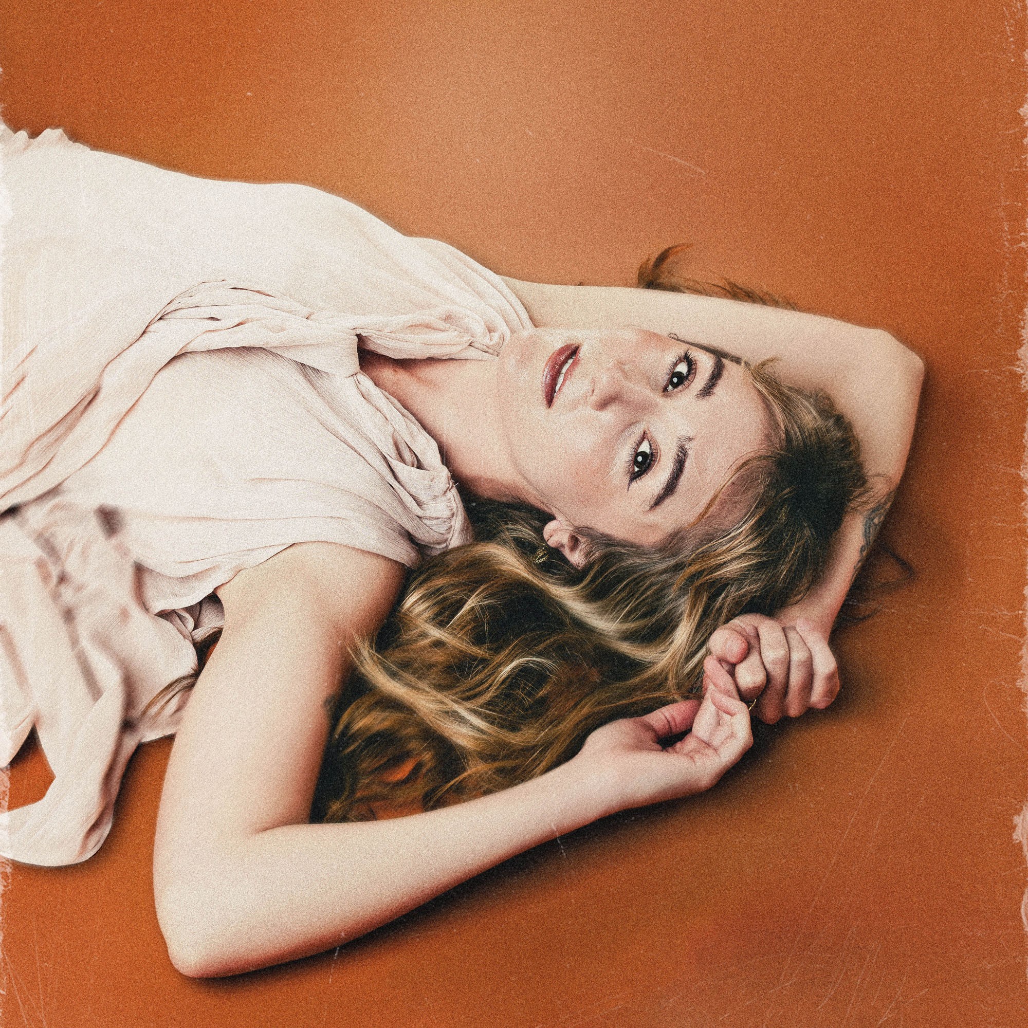 Young woman with long hair lying on an orange surface, resting her head on her arm, looking at the camera.