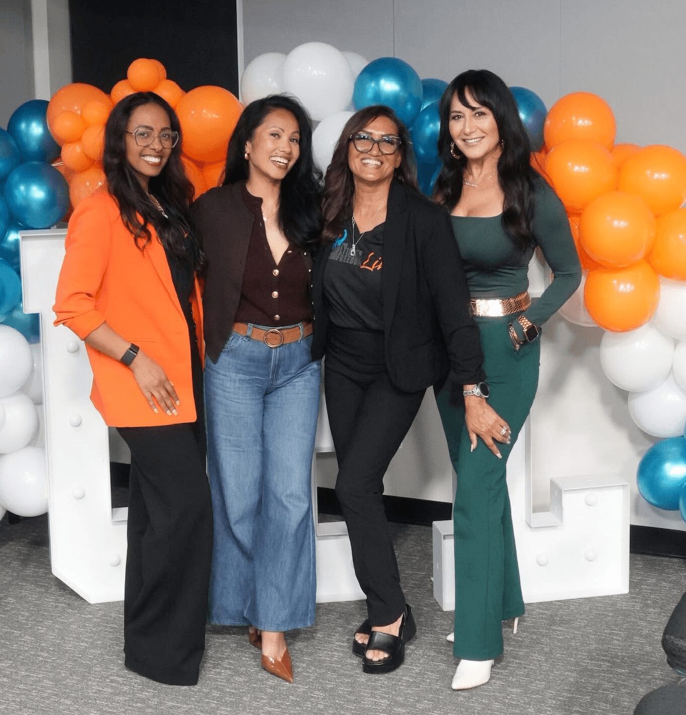 Four women standing together in front of colorful balloon decorations, smiling and posing for the photo.