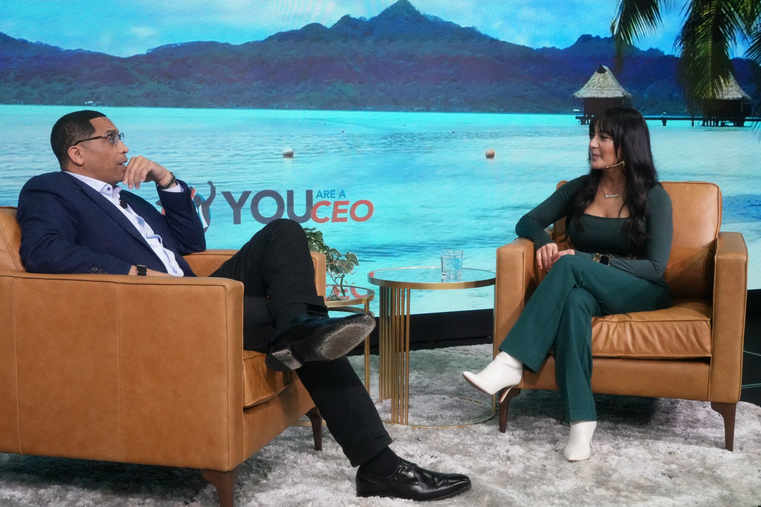 Two people sit on chairs facing each other in a studio with a tropical beach background, discussing.