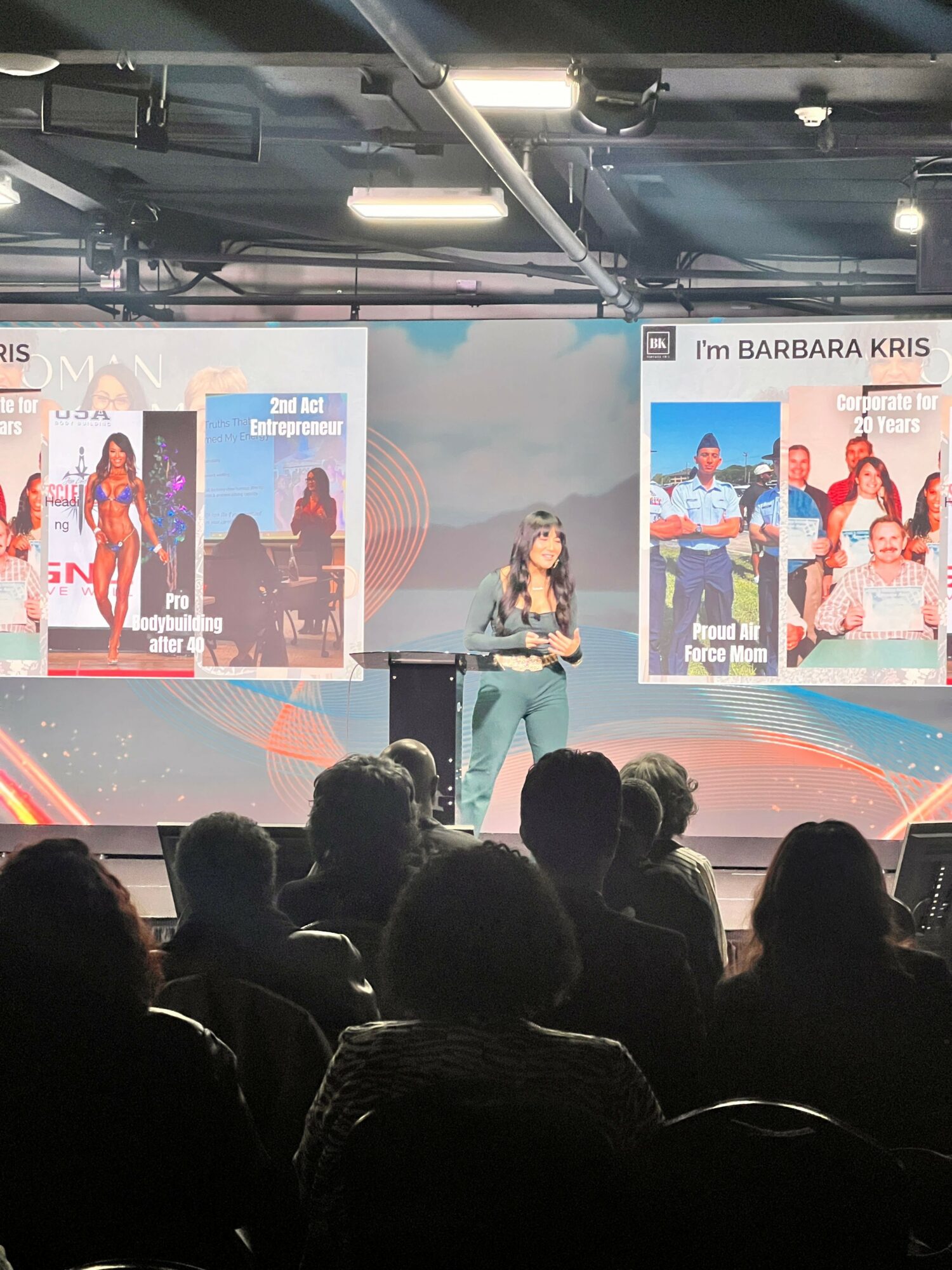 Woman speaking on stage with audience in foreground, large screens displaying images and text behind her.