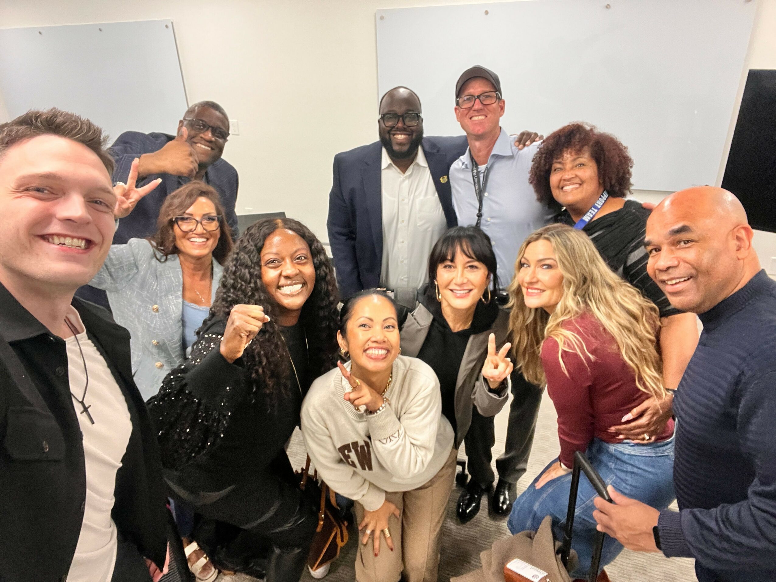 Group of ten diverse people smiling and posing together indoors, some making peace signs, in front of whiteboards.