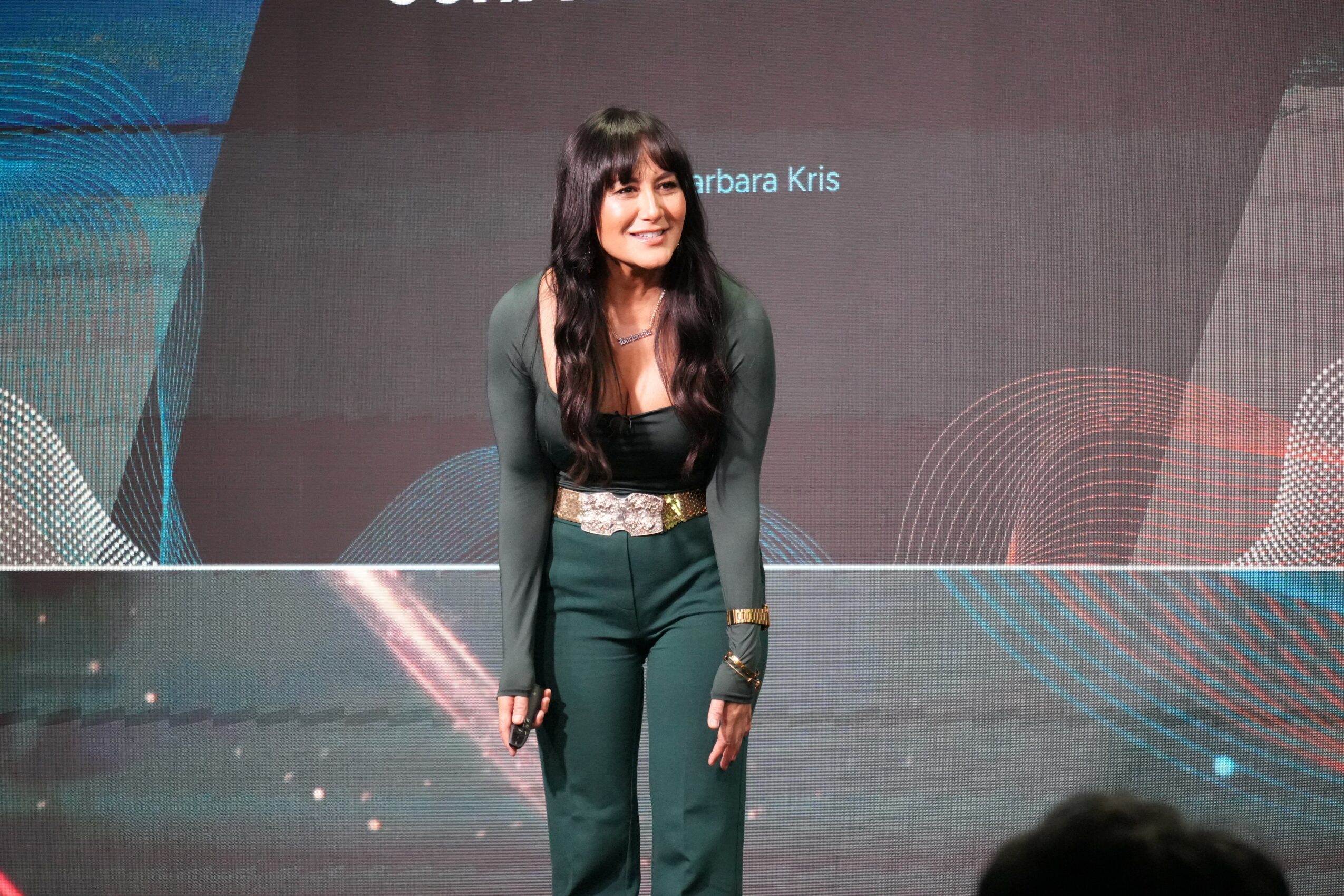 Woman with long dark hair standing on stage, smiling, wearing a dark green outfit with a patterned belt, background with text.