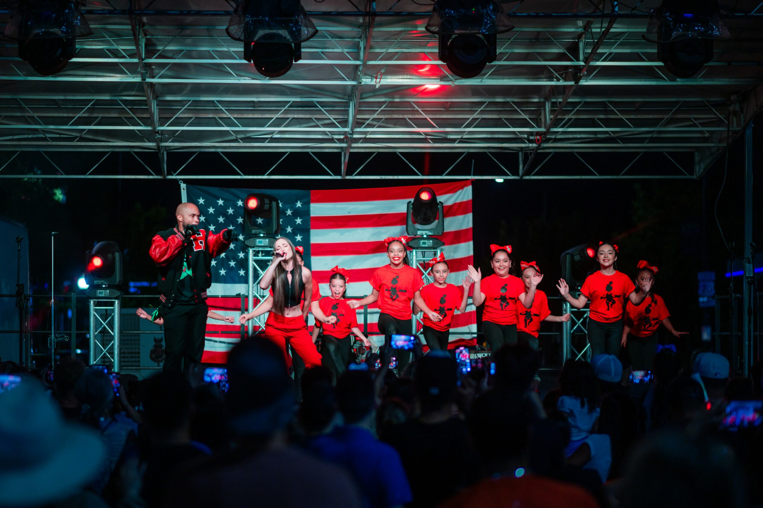 Group of performers on stage with American flag backdrop, singing and dancing, audience in foreground.