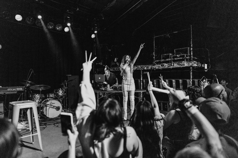 Performer on stage with microphone, audience raising hands, dark background with stage lights, musical instruments visible.