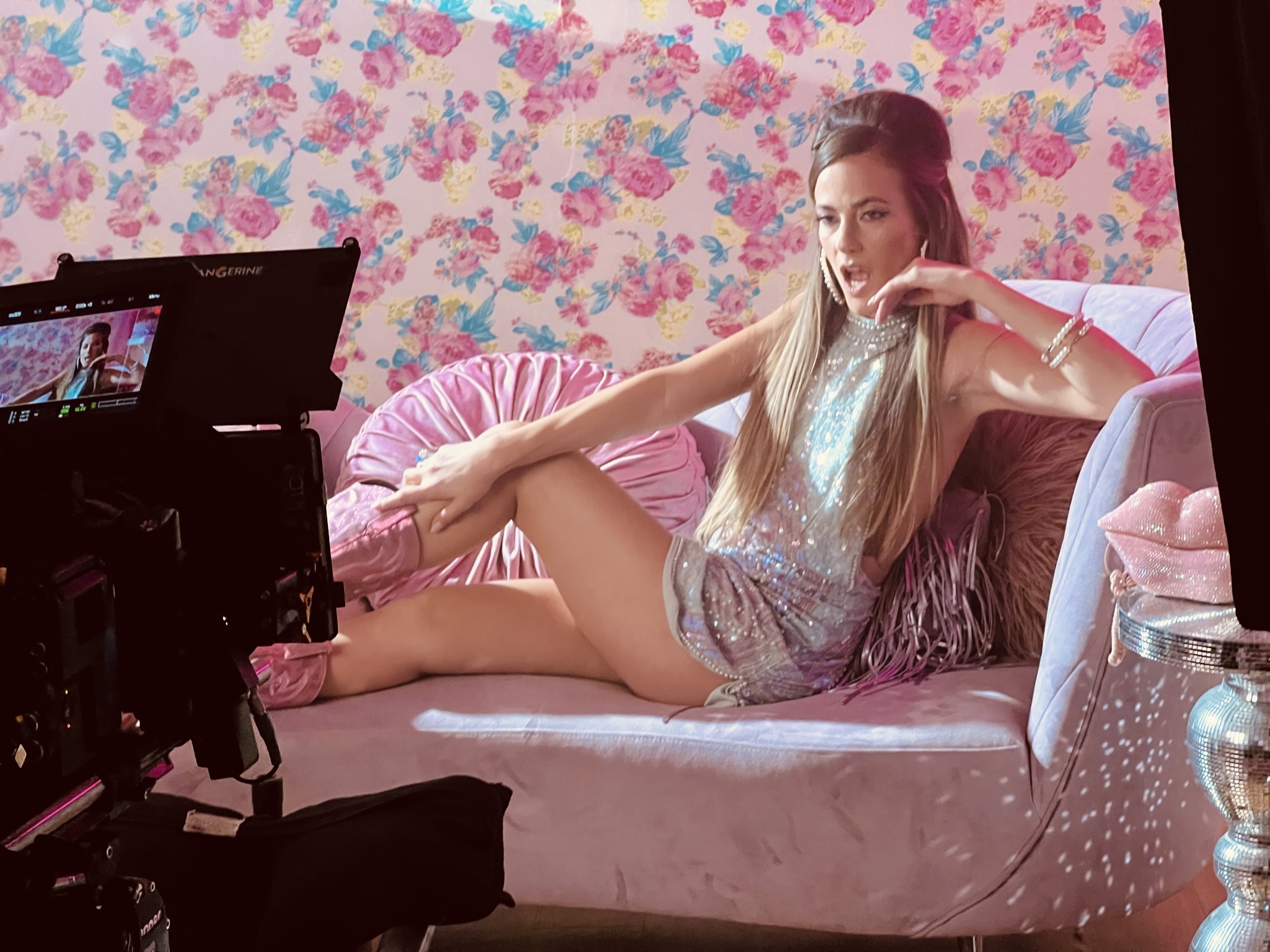 Woman with long hair lying on a pink bed with floral wallpaper background, wearing a shiny dress, in a photoshoot setting.