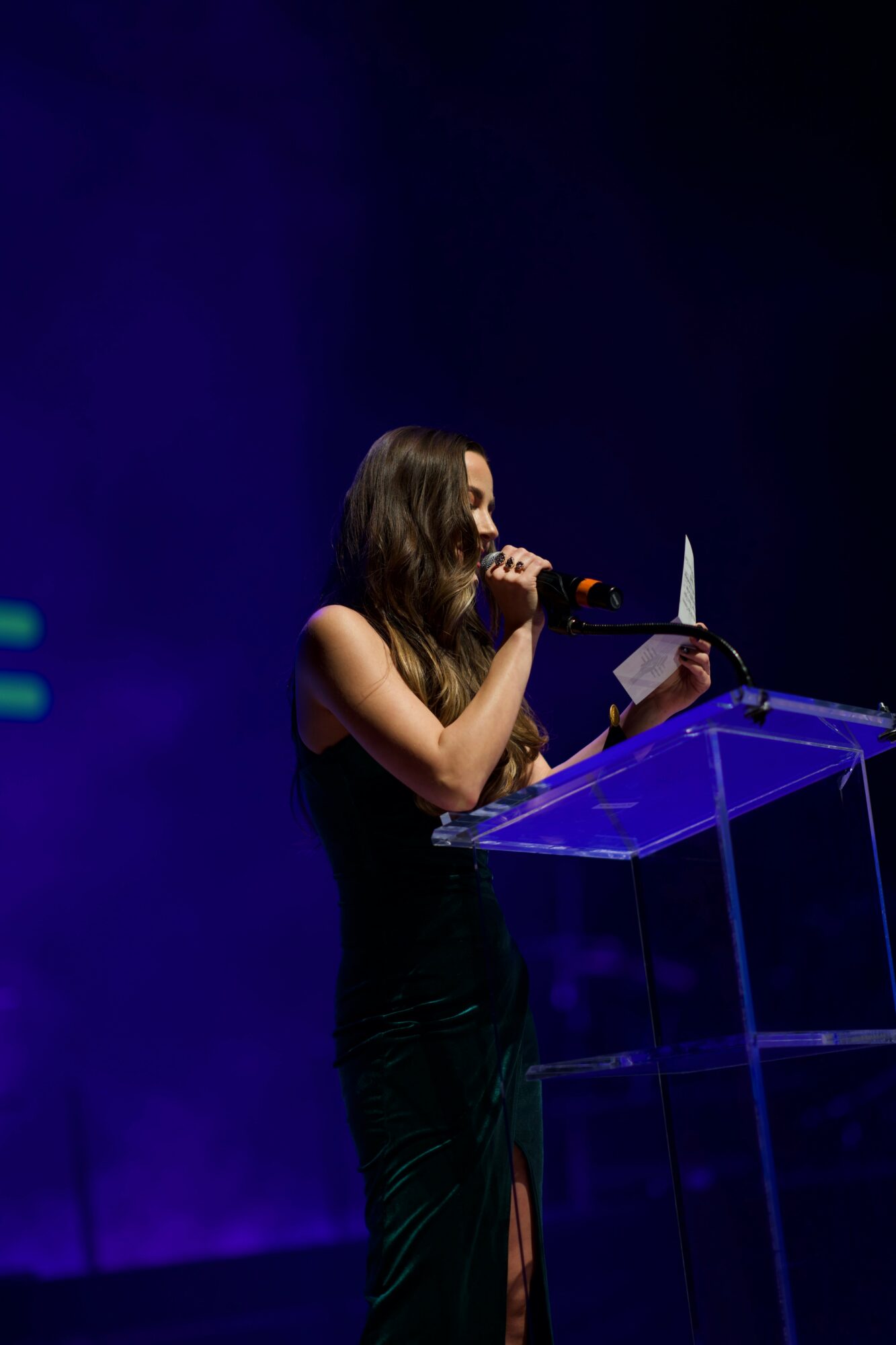 Woman speaking at a transparent podium on stage with dark background.