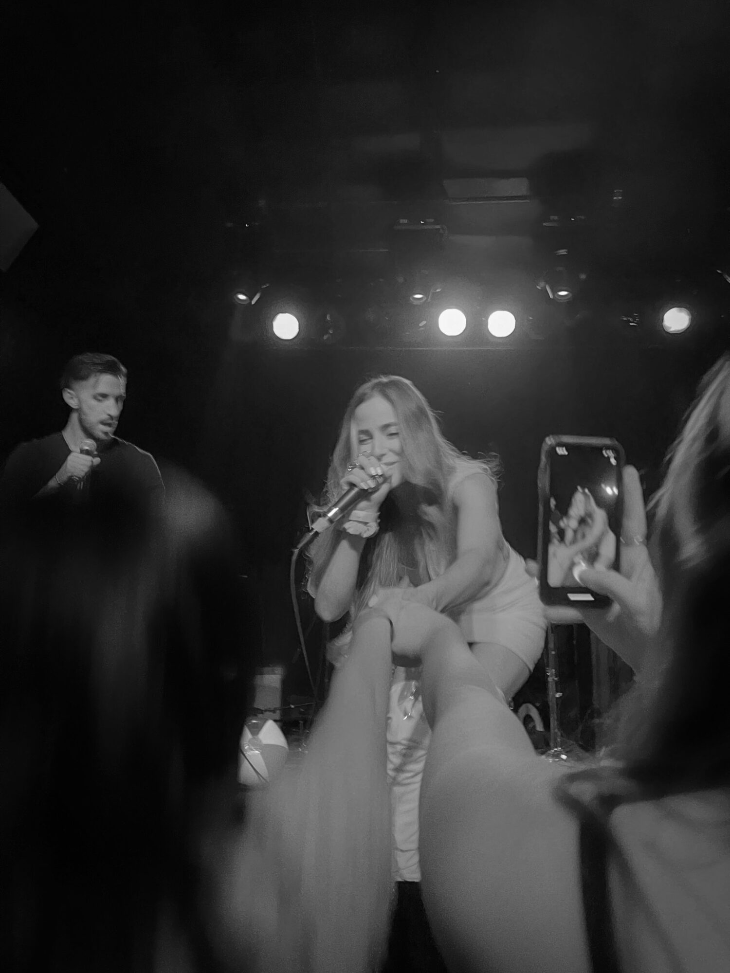 Performer singing on stage with audience members taking photos, stage lights overhead, dark background.