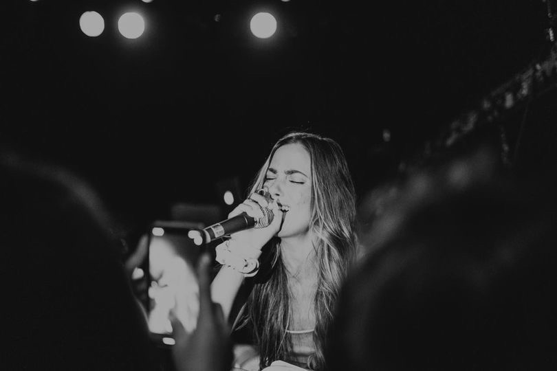 Woman singing into microphone on stage with lights above, audience in foreground, black and white image.