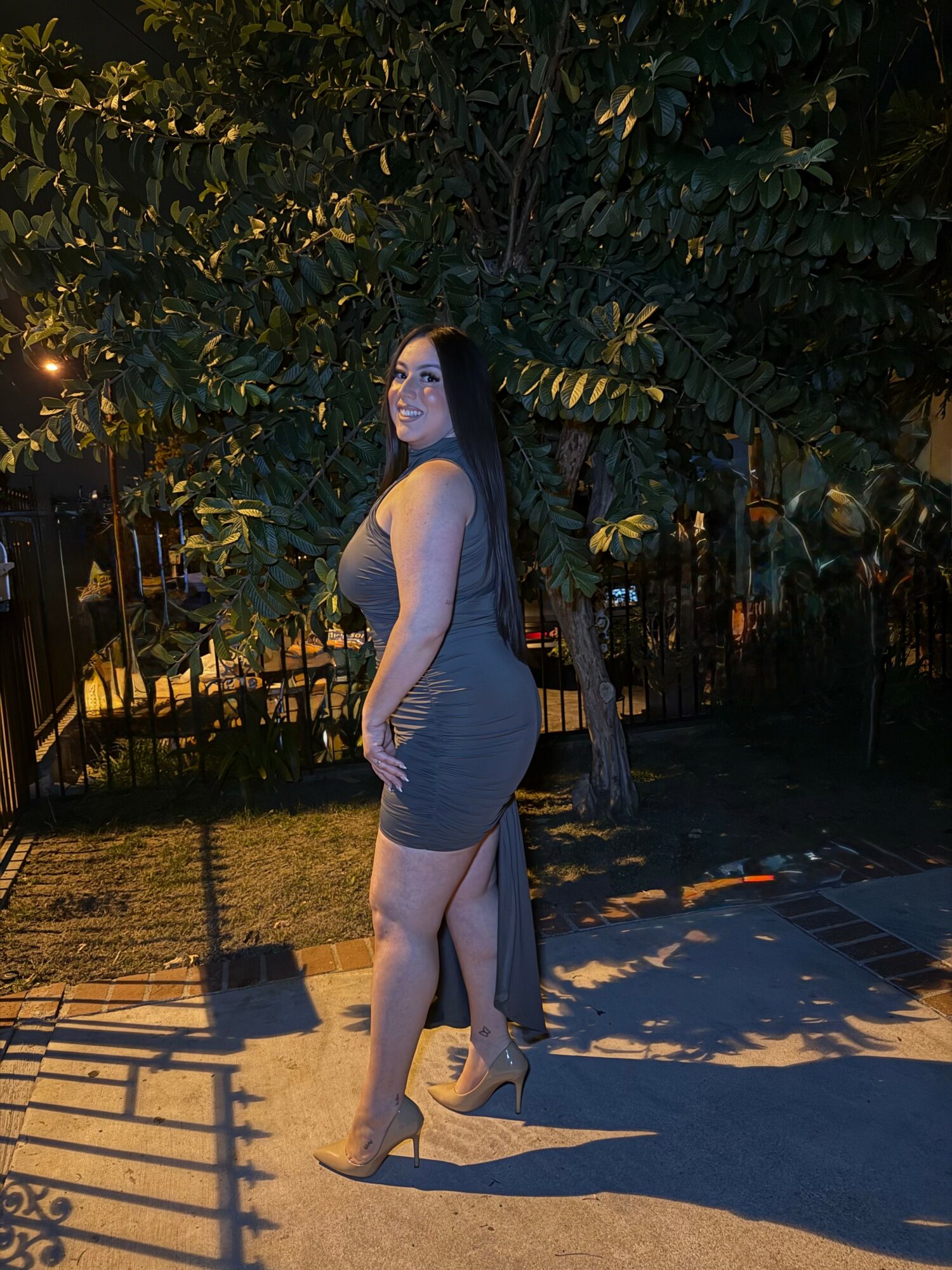 Woman in a gray dress standing outdoors at night with trees and shadows in background.