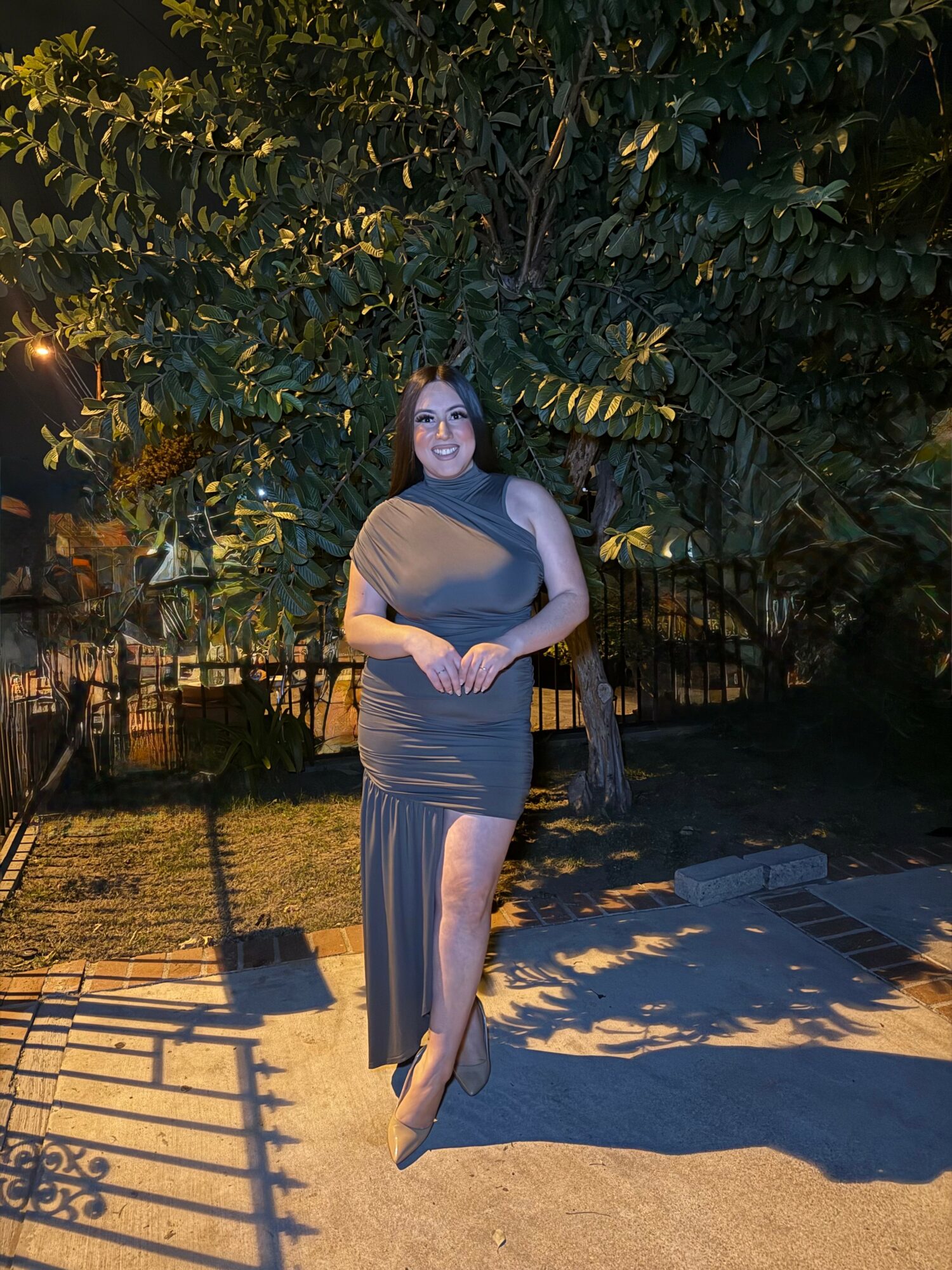 Woman in a dark dress standing outdoors at night with trees and a fence in background.