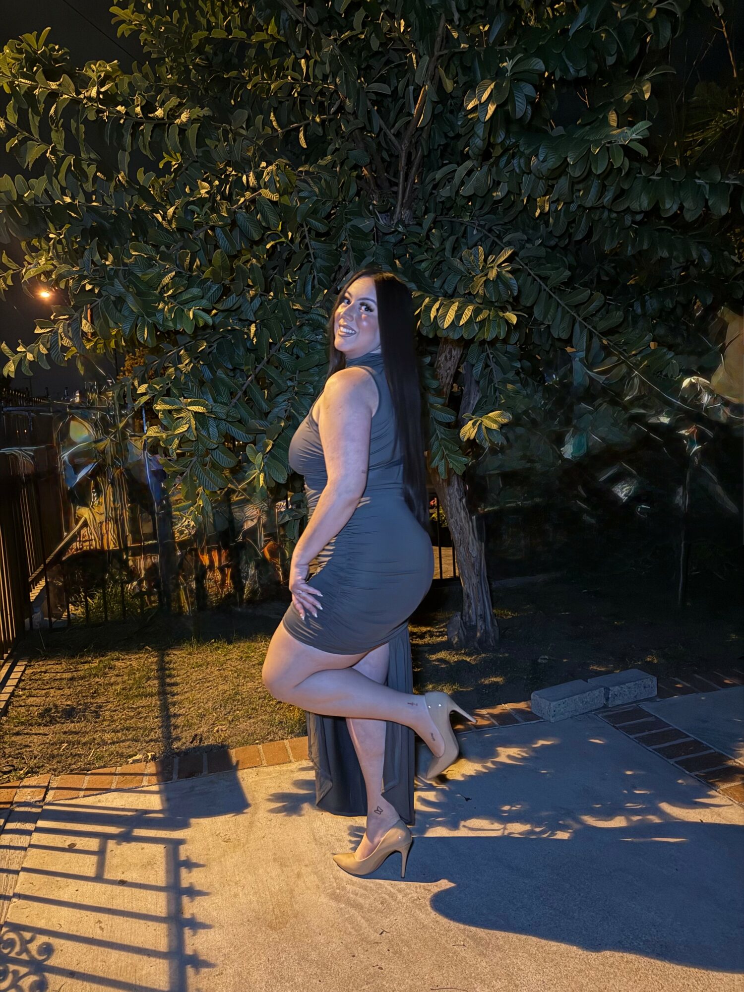 Woman in a gray dress standing outdoors at night, surrounded by trees and shadows.